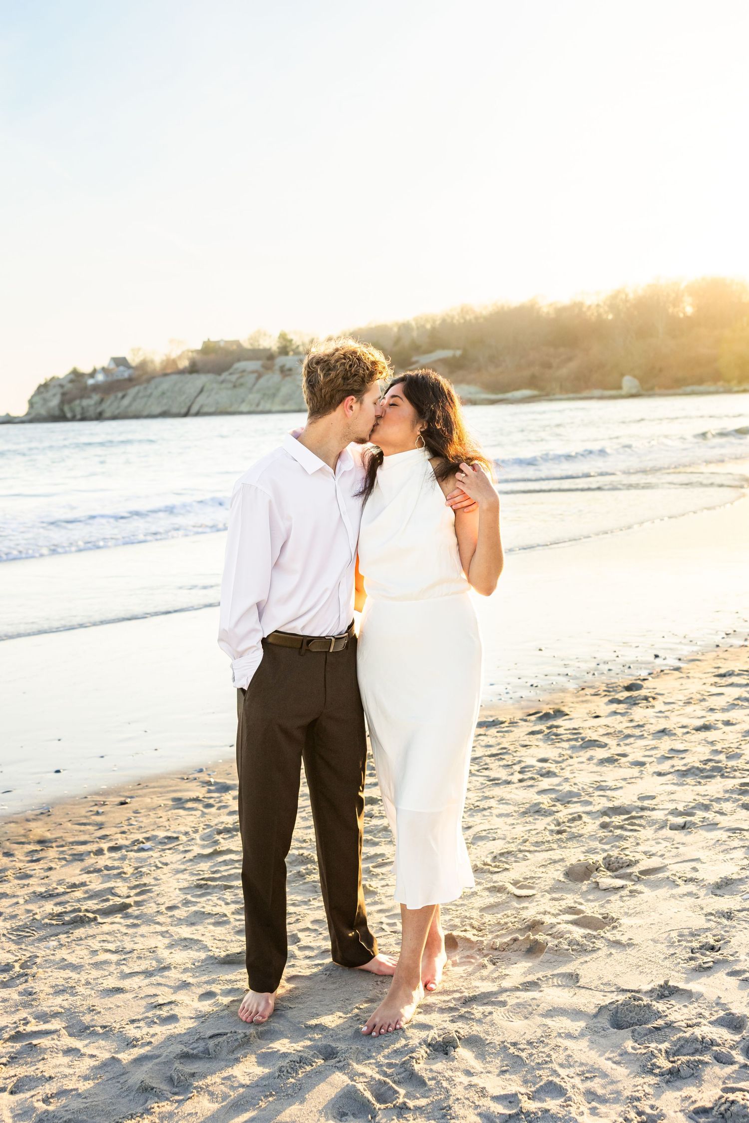 A romantic couple shares a tender moment on a sunlit beach at sunset during their seaside photoshoot.