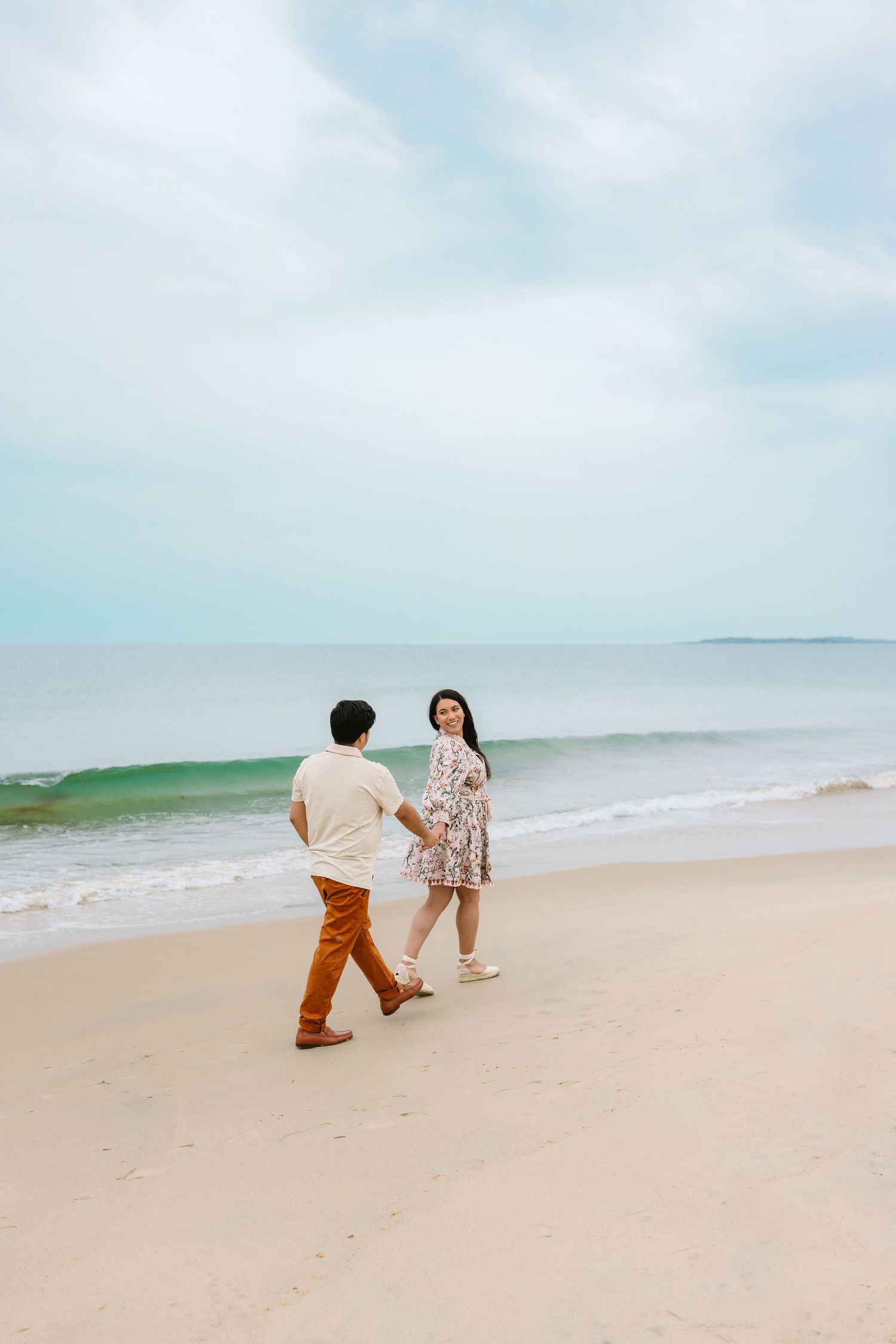 Couple walking hand in hand along a sandy beach with ocean waves behind them.