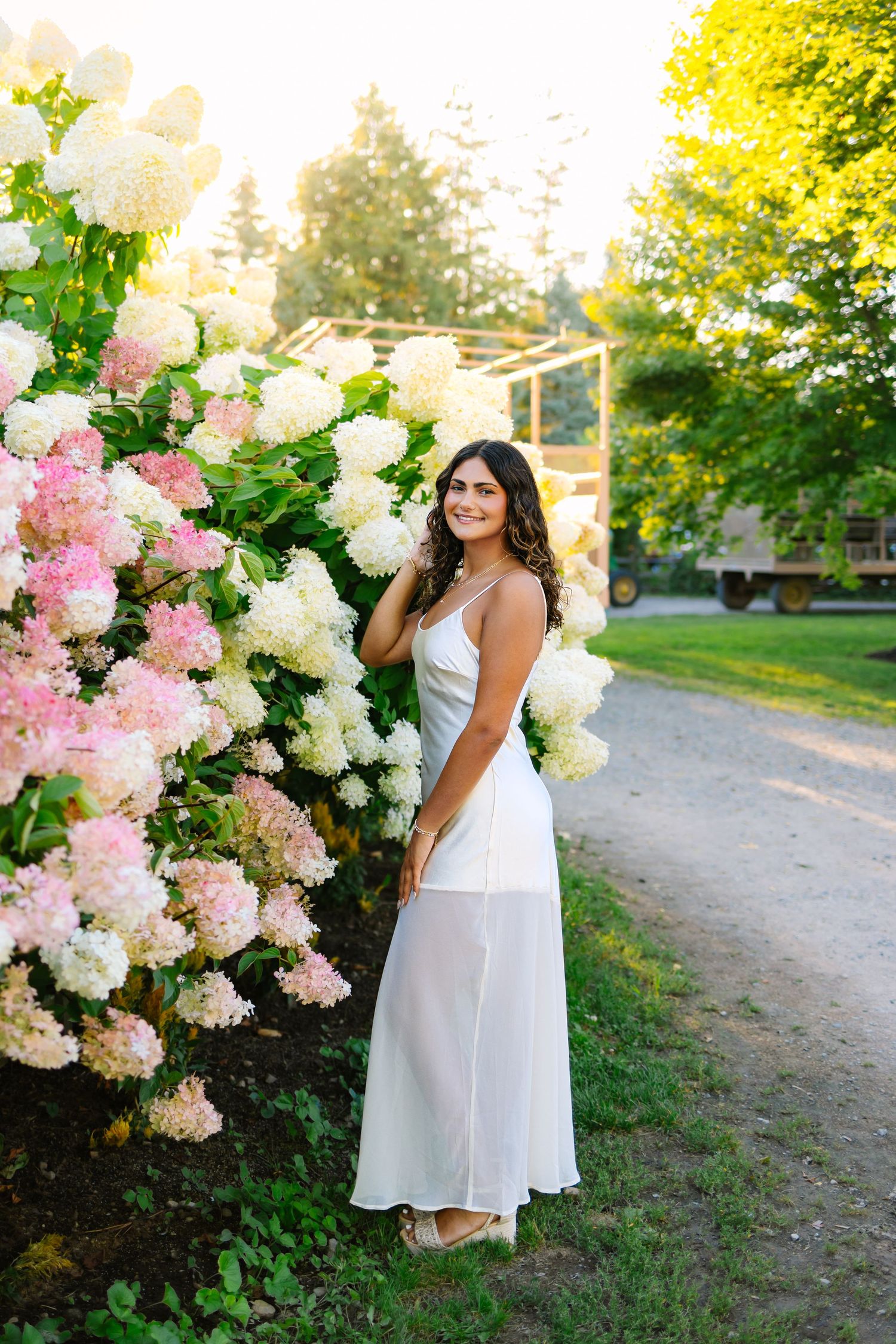 A smiling brunette woman in a white slip dress poses beside blooming pink and white hydrangeas at sunset.