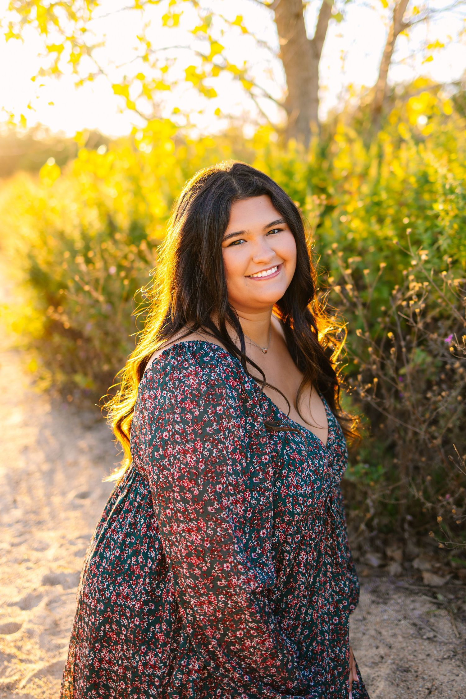 Smiling young woman in floral dress on sandy path surrounded by golden foliage.