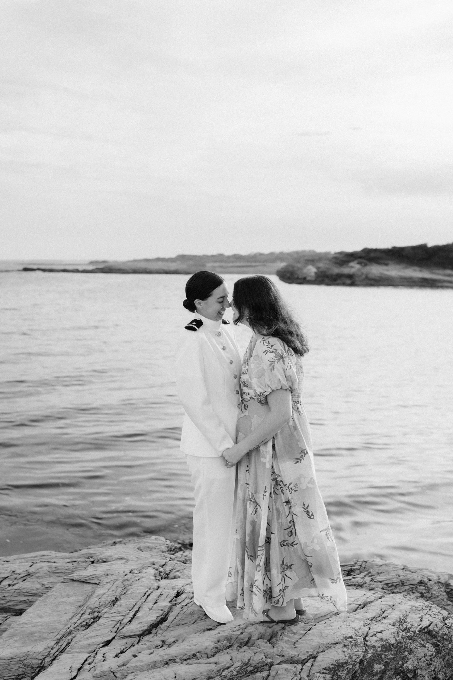 A couple shares an intimate moment on rocky shore in black and white coastal photography.
