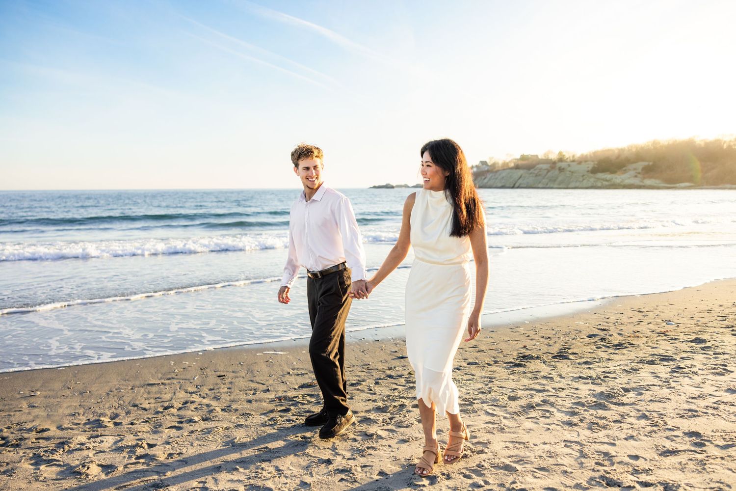 A couple walks hand in hand along a sunny beach shoreline during golden hour.
