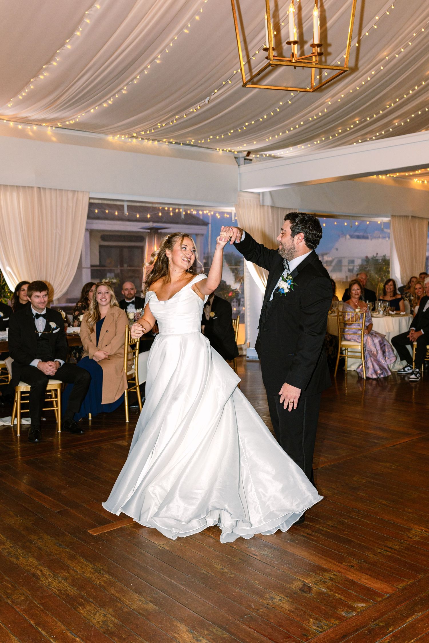 A couple shares a romantic dance on a wooden floor under draped fabric and golden chandeliers at an elegant wedding reception.