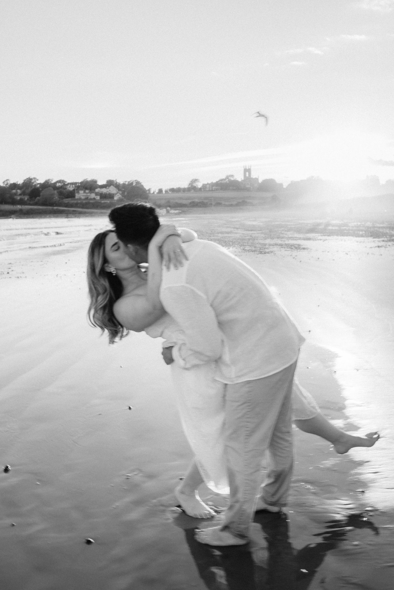 Black and white romantic beach photo of couple kissing, man dipping woman near ocean shoreline.