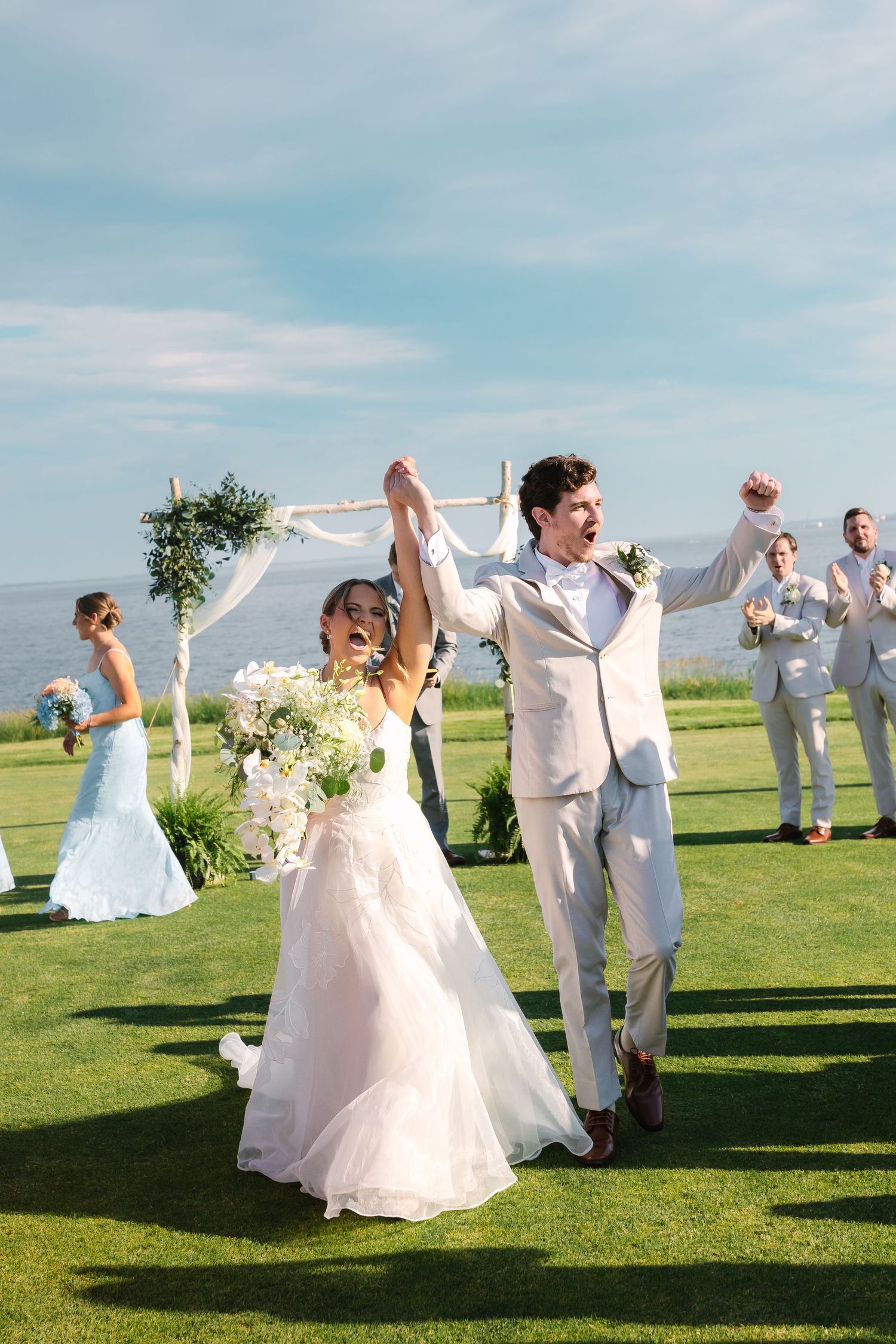 A joyful bride and groom raise hands in celebration at an outdoor waterfront wedding ceremony.