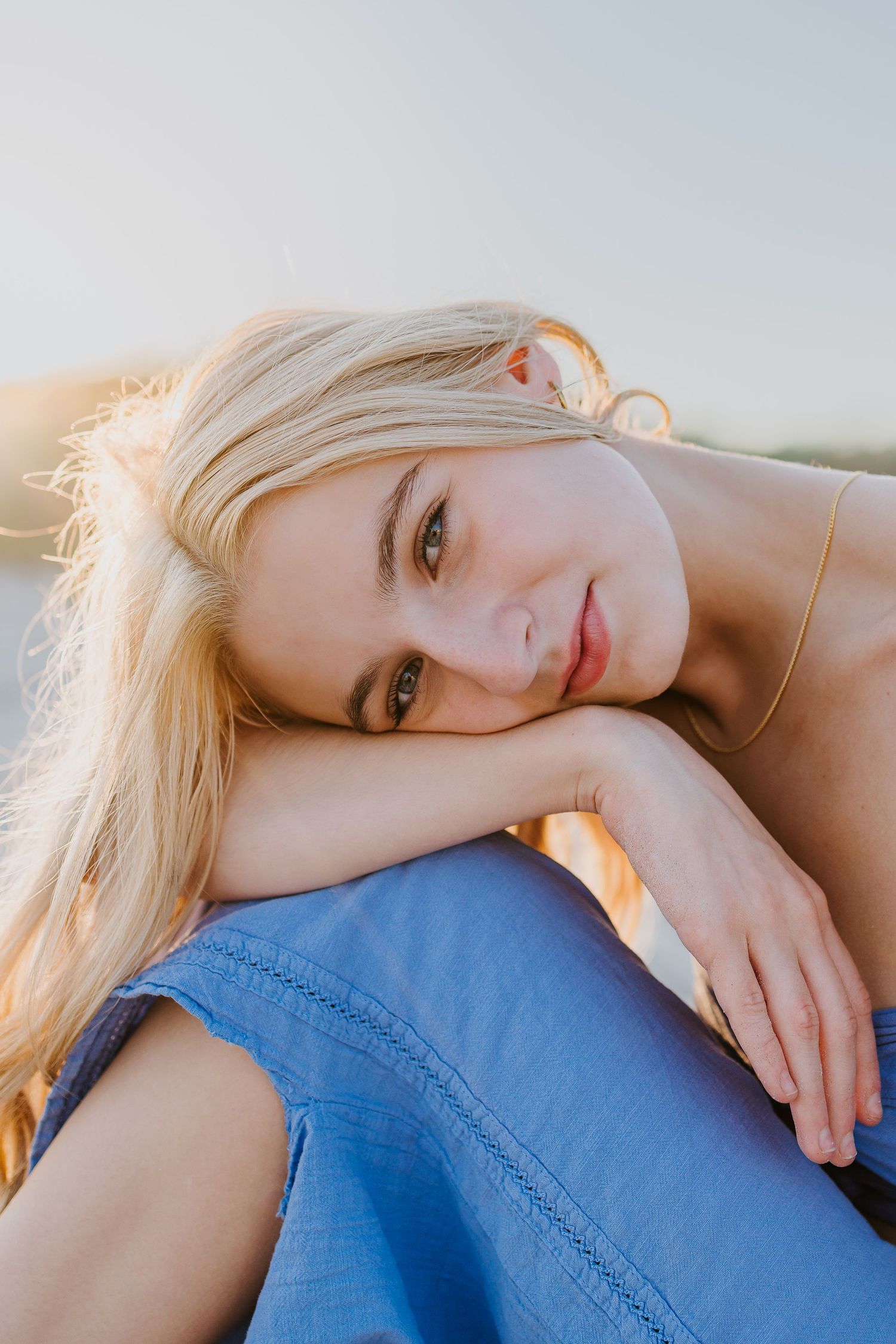 Close-up portrait of a blonde woman resting her chin on her knees in golden sunset light.