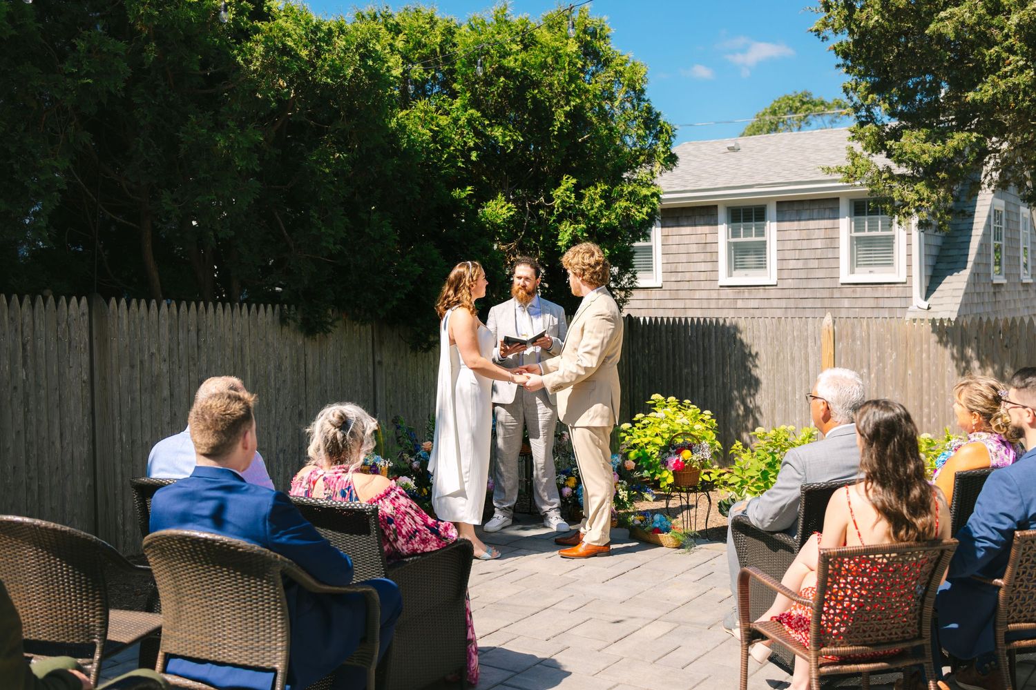 Outdoor backyard wedding ceremony with guests seated watching couple exchange vows on sunny summer day.