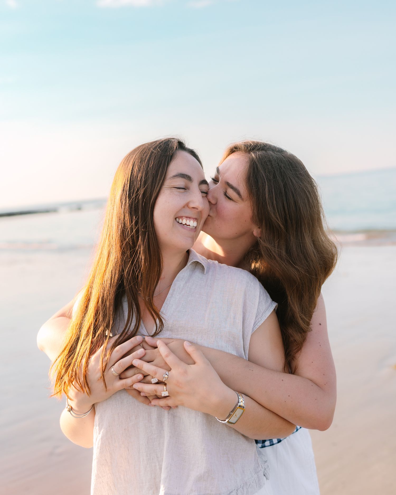 Newly engaged couple embracing on beach, one woman smiling as her partner kisses her cheek.