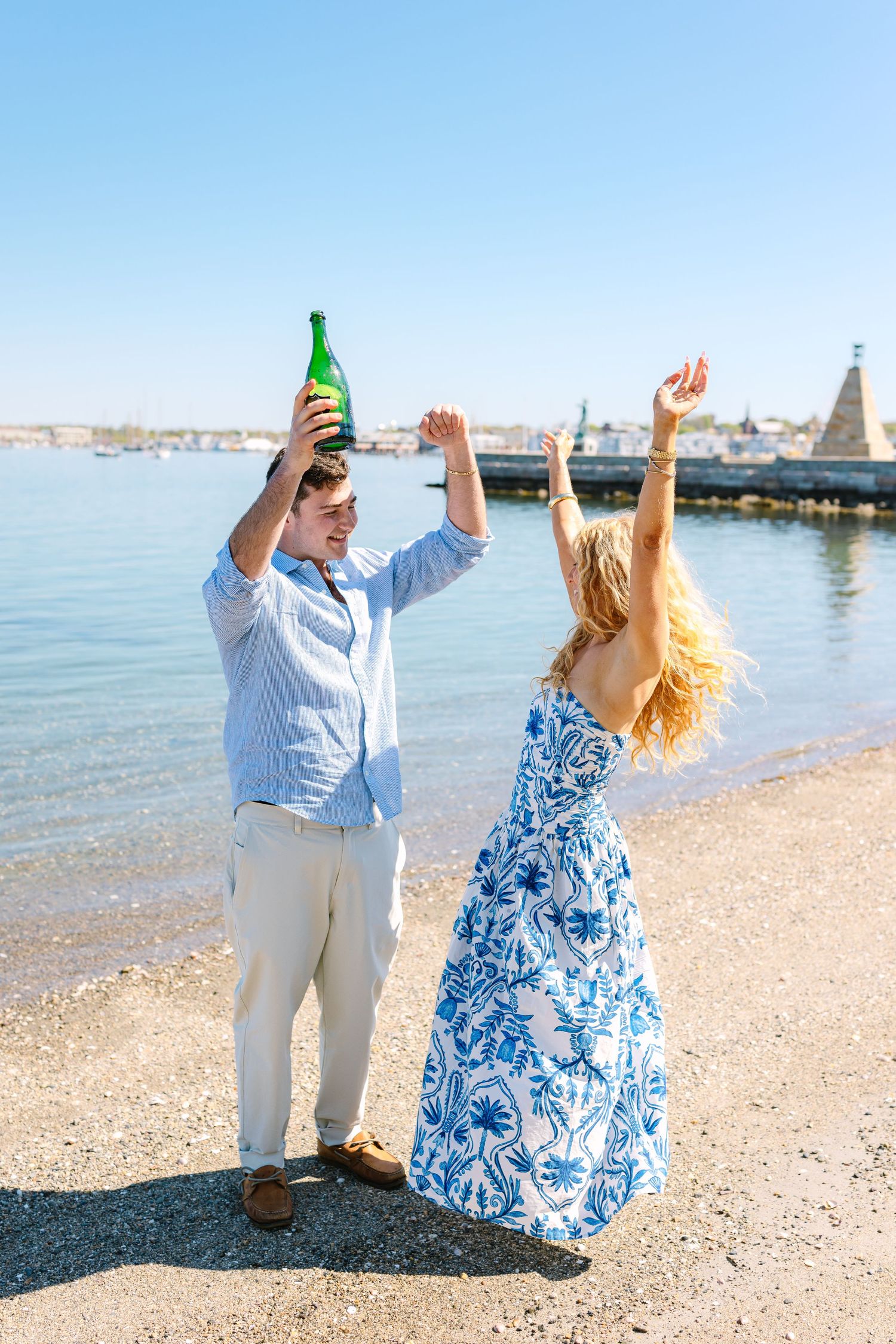 Excited couple celebrating on beach with champagne bottle raised, woman with arms up in joy.
