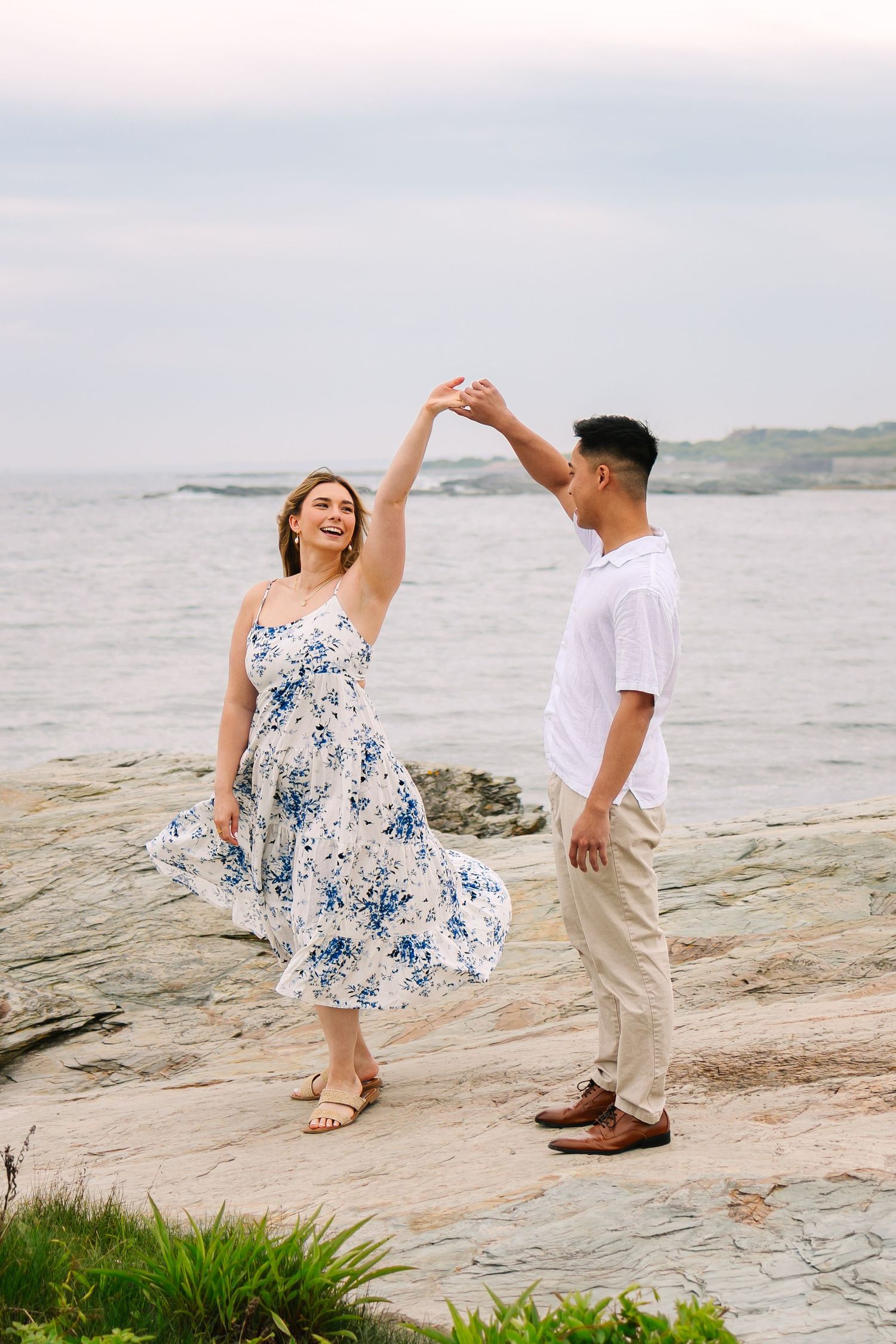 Couple dancing on rocky coastal shoreline for engagement photos, woman in floral blue dress being twirled by man in Newport Rhode Island