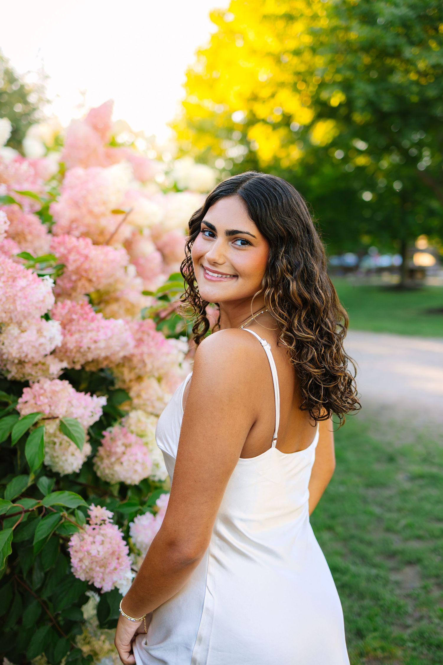 Young woman in white dress smiling over shoulder beside pink hydrangea blooms at golden hour.
