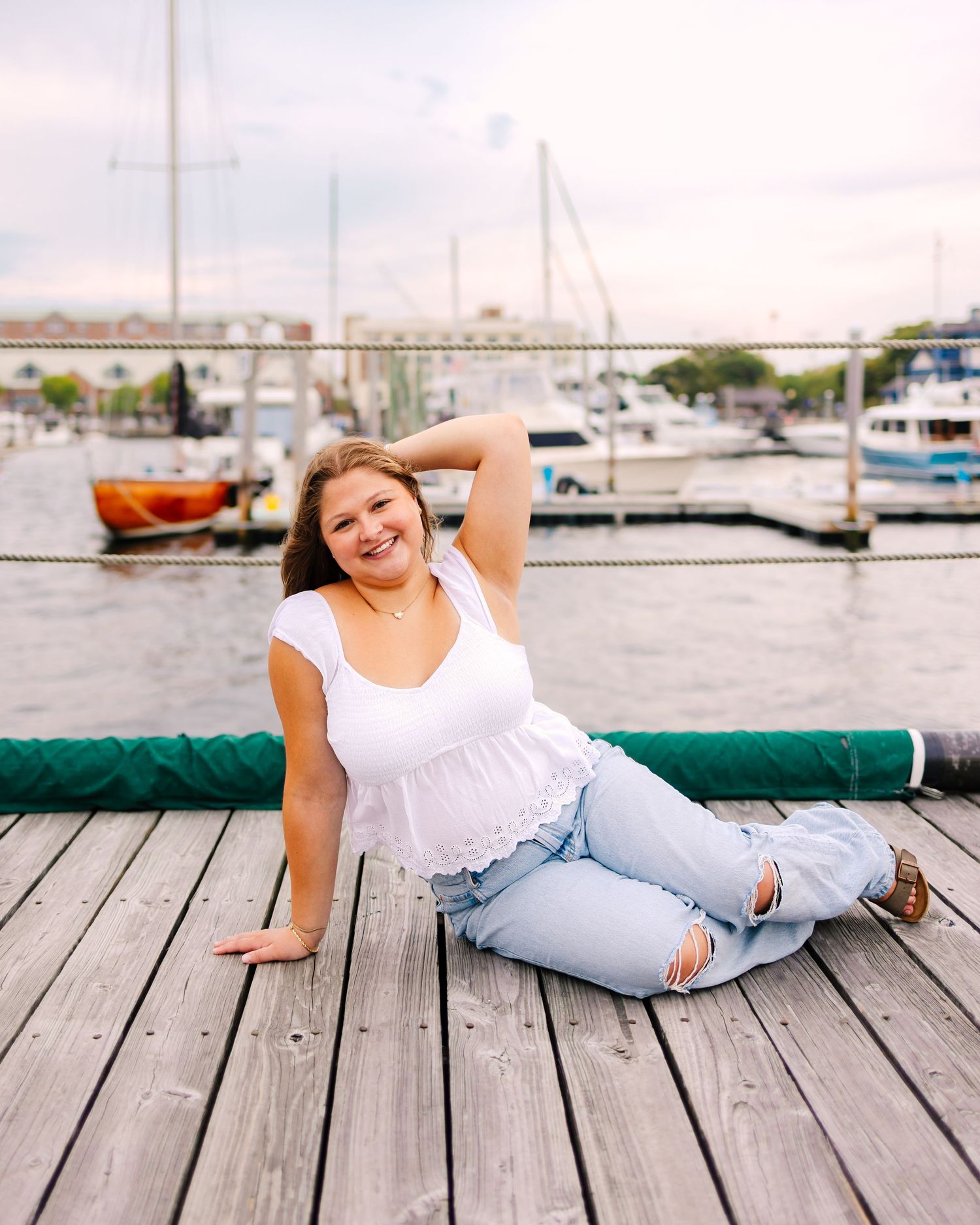 Happy young woman posing on a waterfront dock in white top and ripped jeans with marina boats behind her.