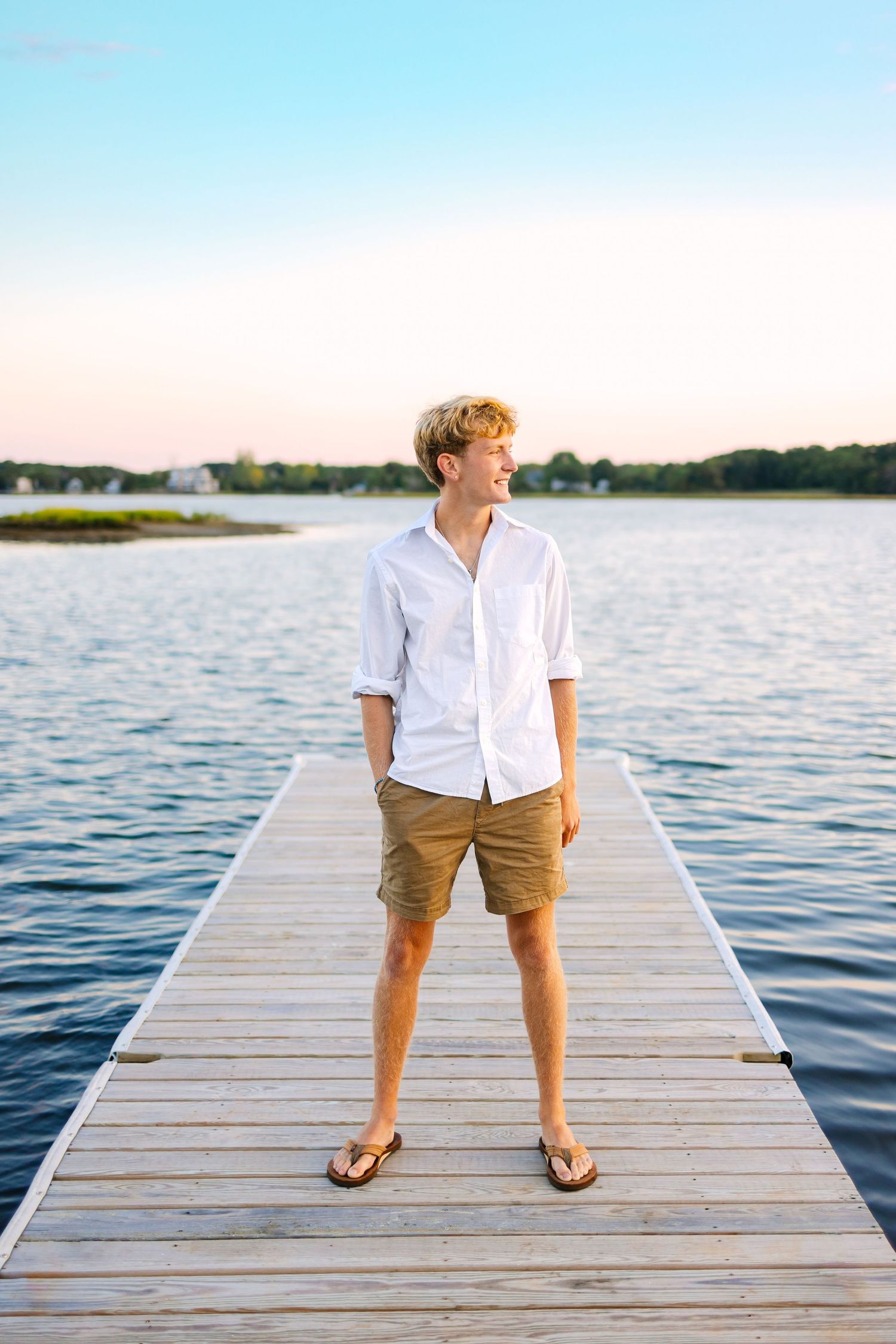 Teen boy in white shirt and khaki shorts stands confidently on wooden dock overlooking serene lake at dusk.