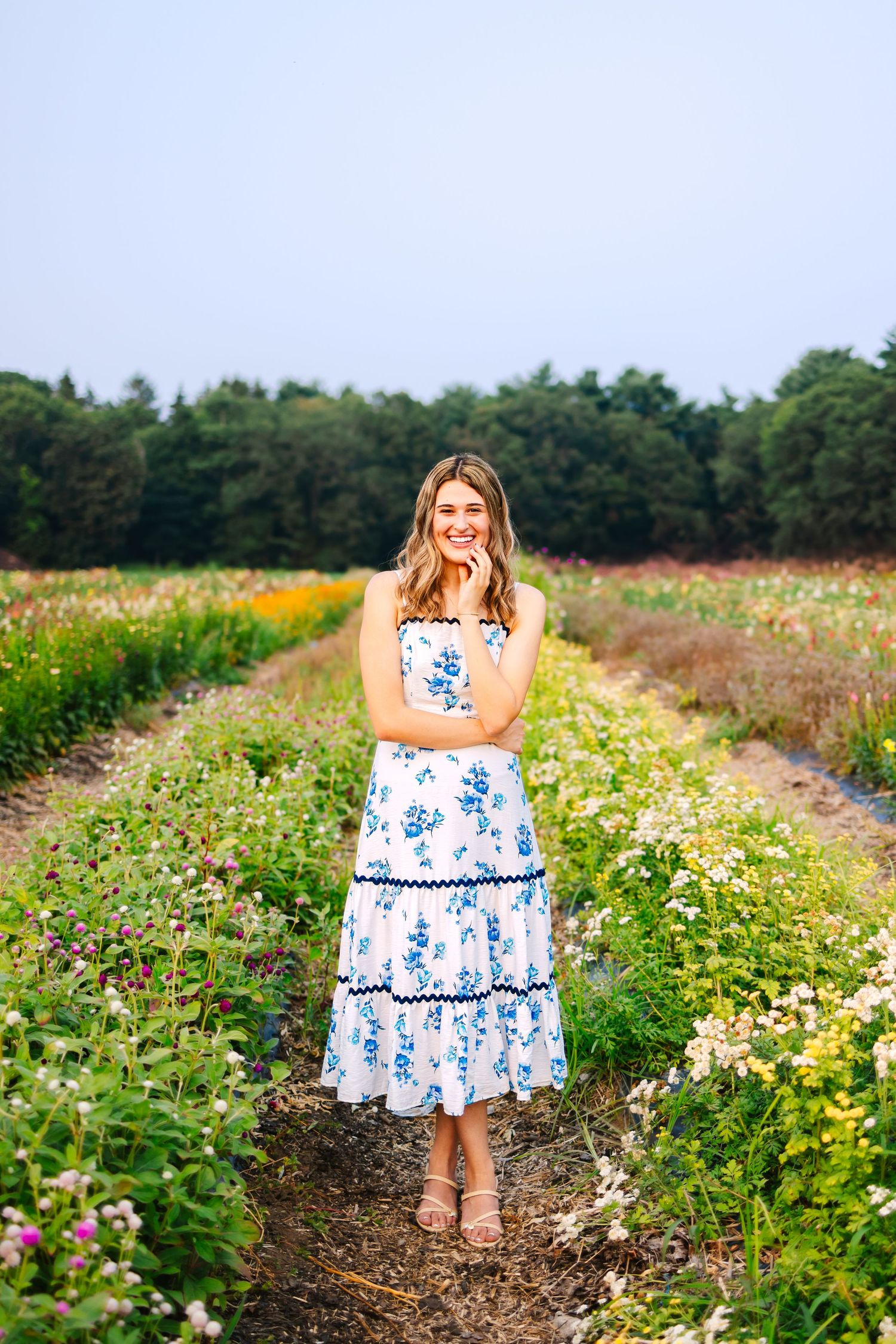 Happy young woman in white blue floral dress standing and smiling in lush colorful flower farm field.