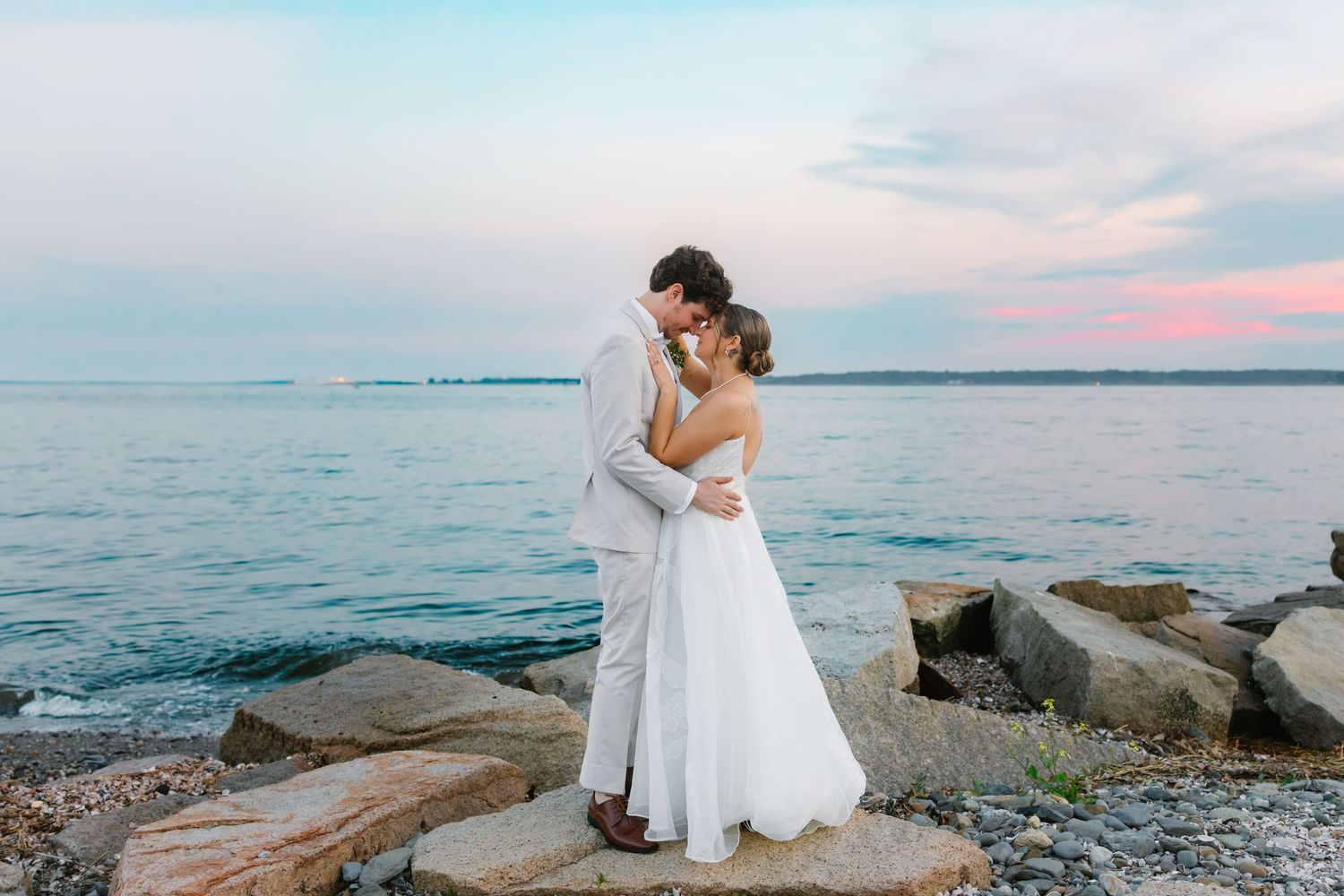 Bride and groom kissing on rocky beach with pastel pink and blue sunset over water.