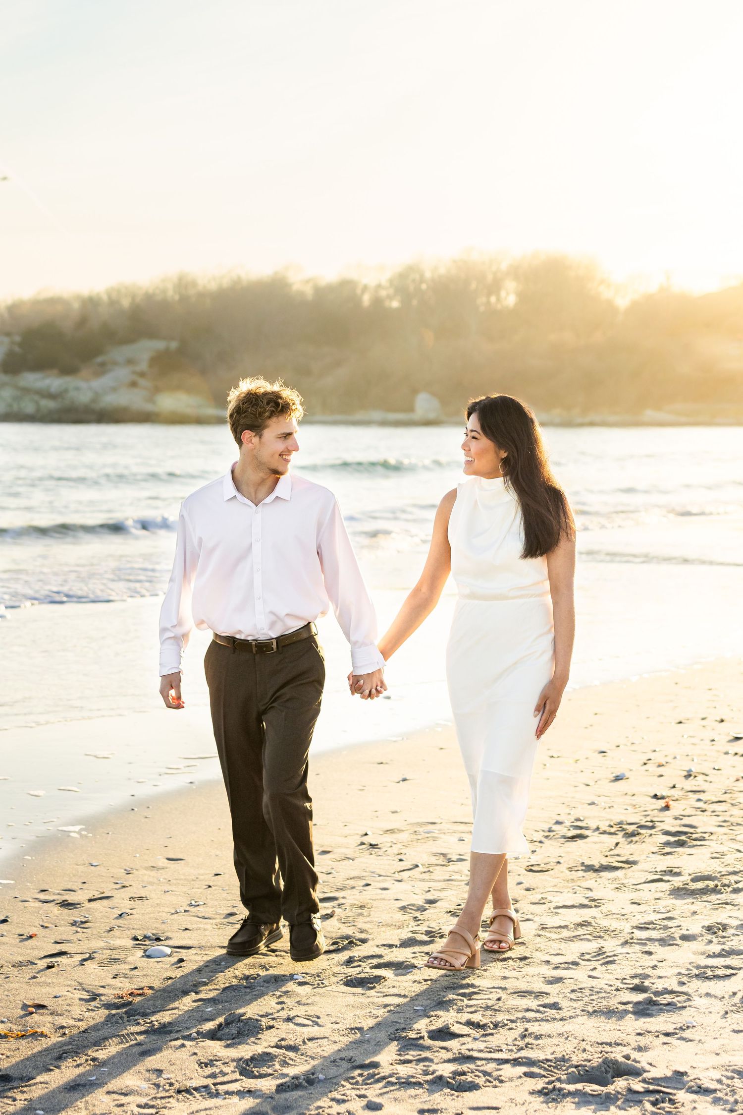 Elegant couple walking hand in hand along sunlit beach shoreline at golden hour sunset.