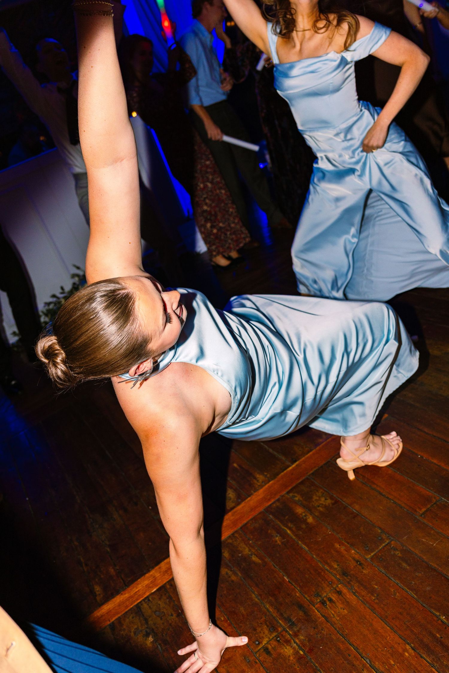 Dancer in light blue dress performs a dramatic breakdance move on a wooden floor at a party venue.