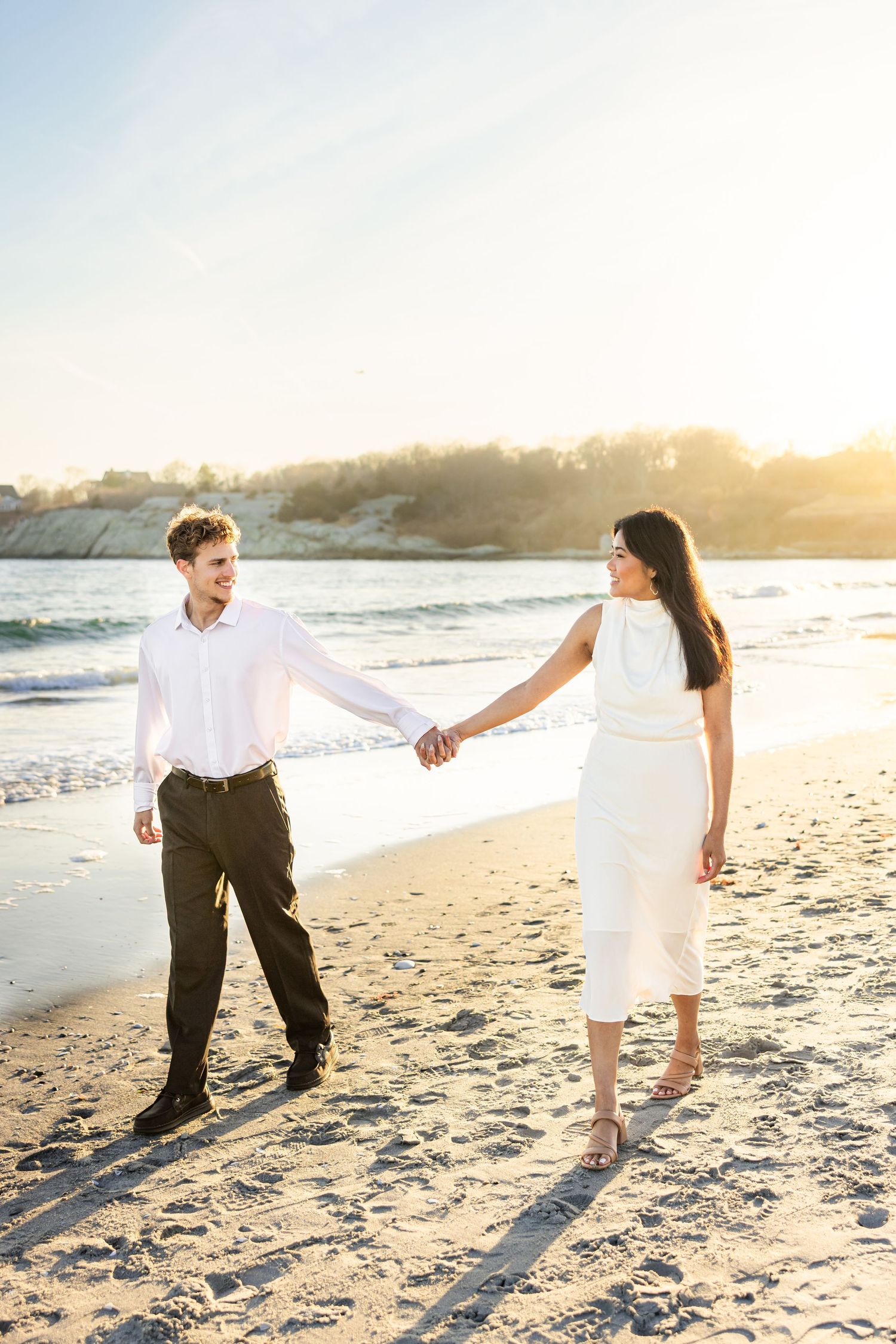 Couple holding hands and walking along a scenic beach at sunset, casting long shadows on the golden sand.