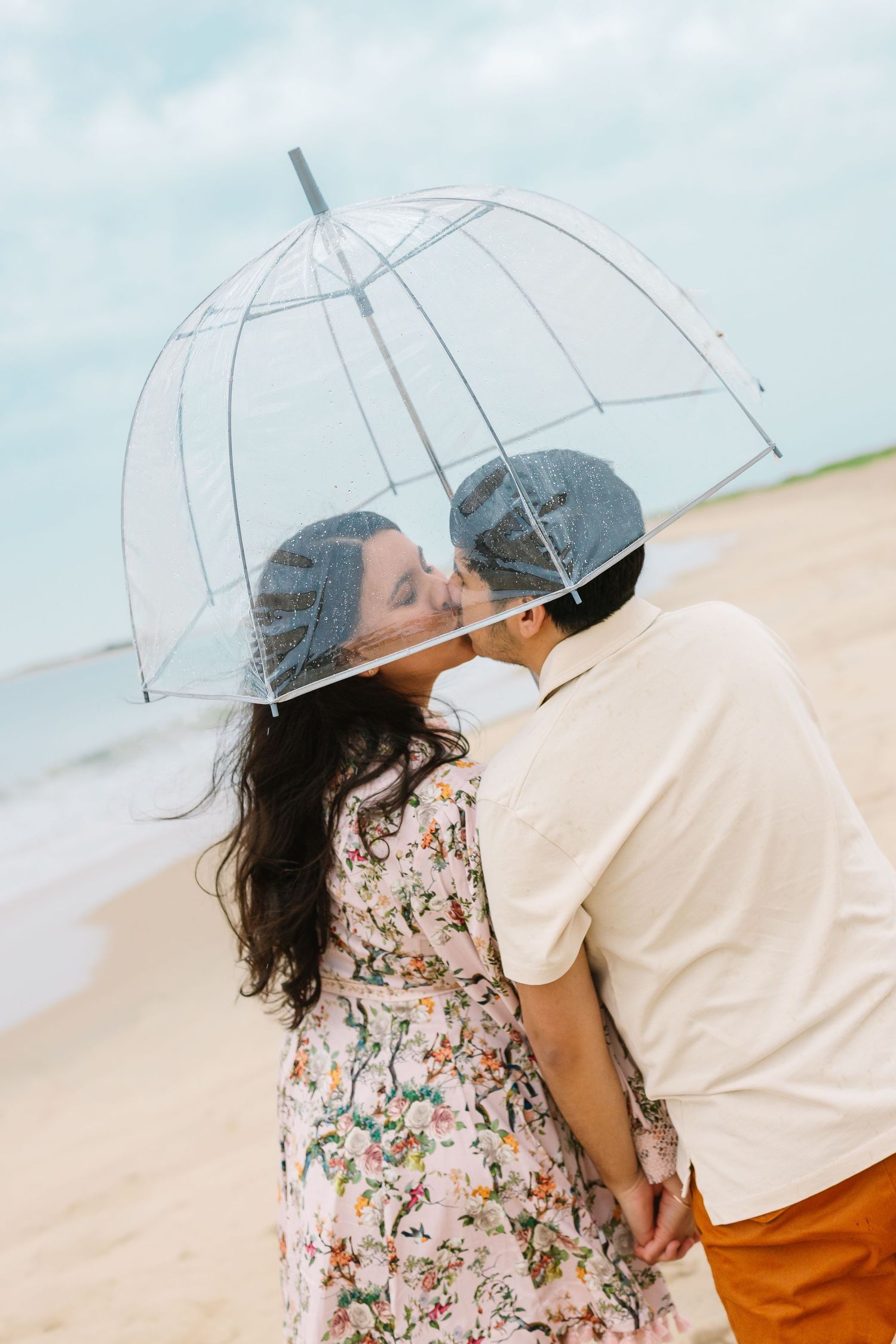 Couple sharing a romantic kiss under a clear bubble umbrella on a beach.