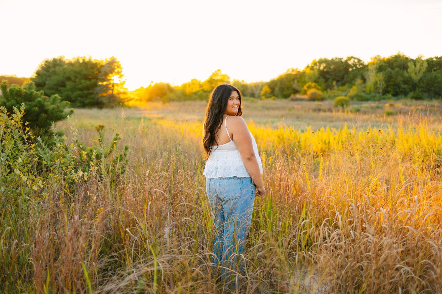 Young woman in white top and jeans standing in tall grass field at sunset.