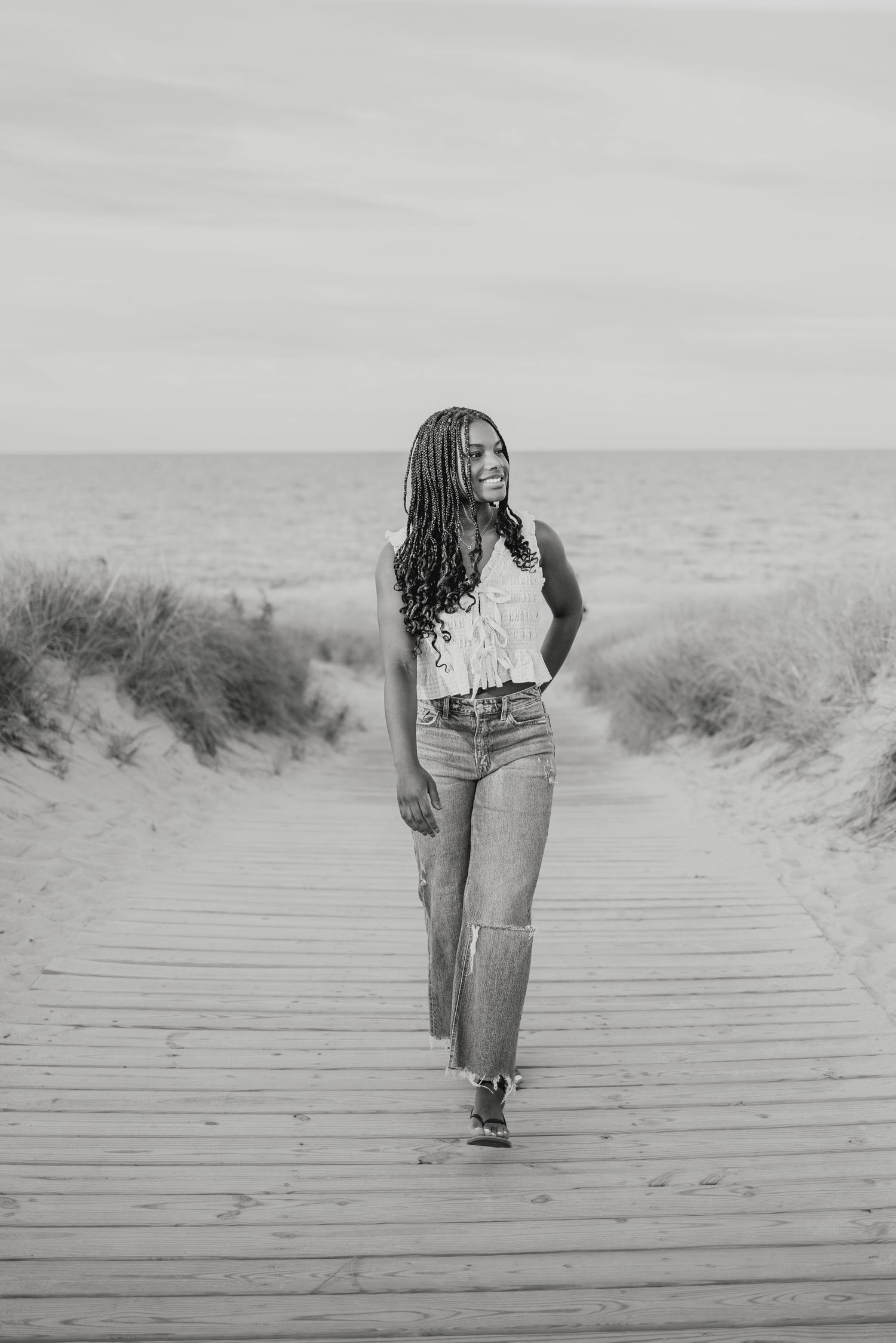 Black and white portrait of young woman with braids walking on wooden beach boardwalk toward the ocean.