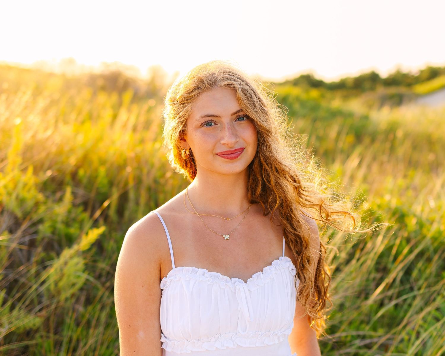Portrait of a young woman with long auburn hair in a white dress standing in sun-kissed beach grass.