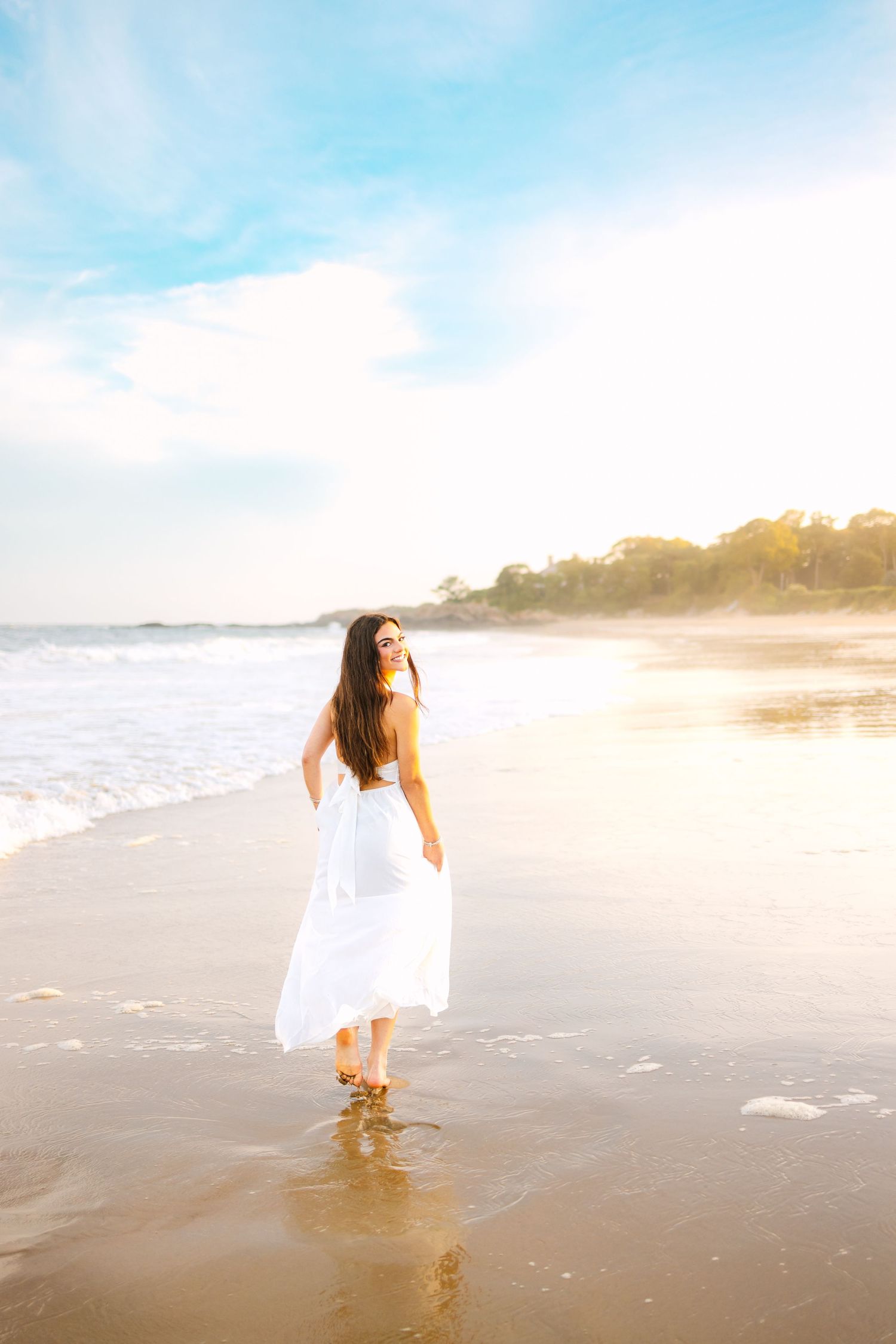 Woman in flowing white dress walking barefoot on wet sandy beach in warm golden sunlight.