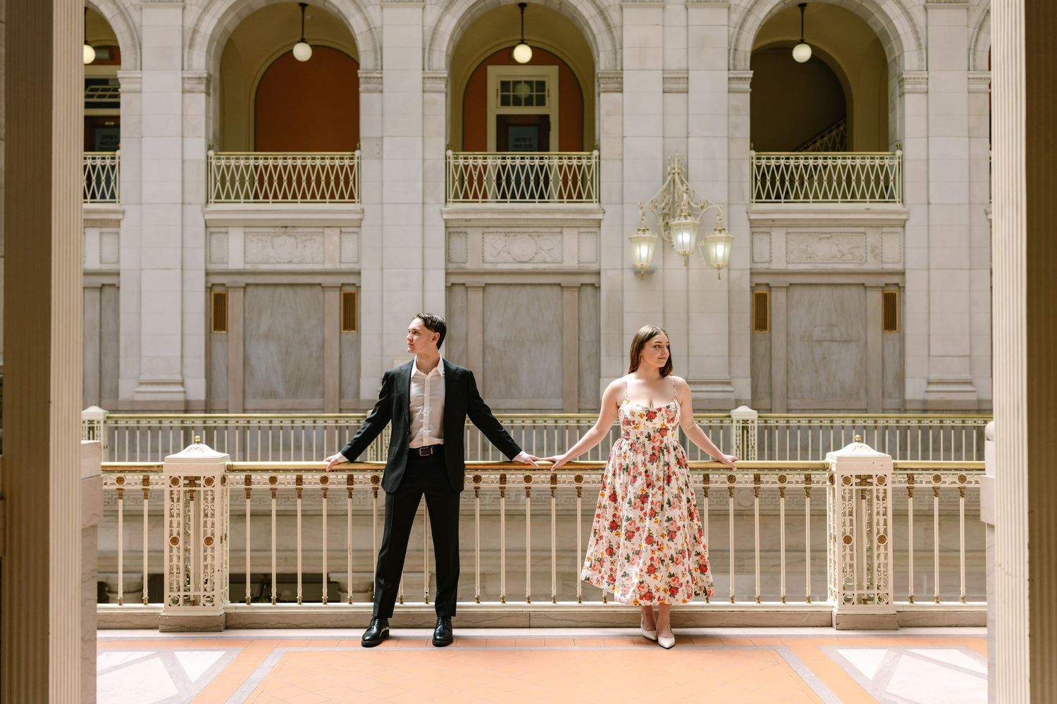 Engaged couple holding hands apart on a balcony inside a grand historic building with arched columns.