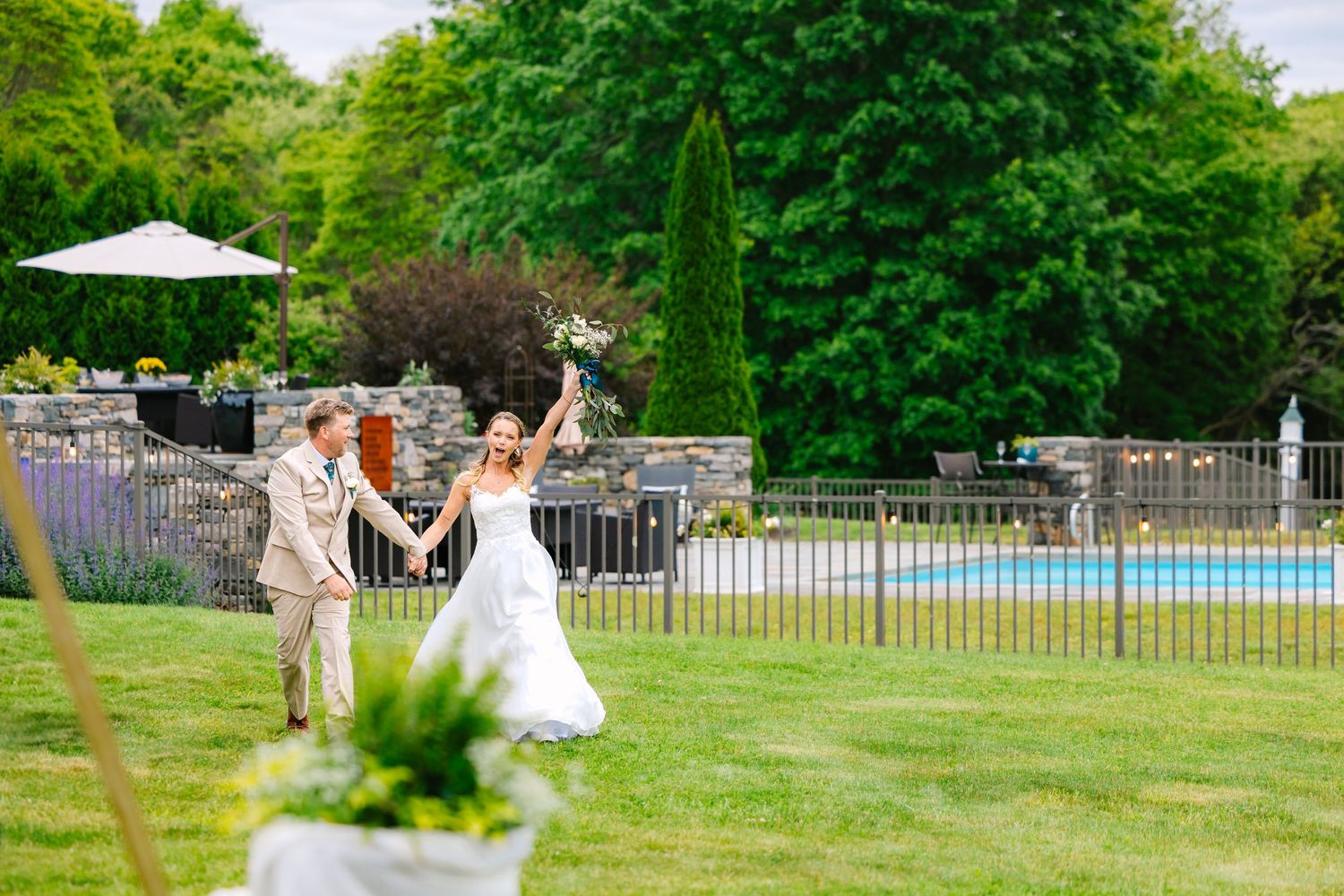 Celebratory moment between newlyweds dancing on green lawn near pool area with white fence and patio umbrellas.