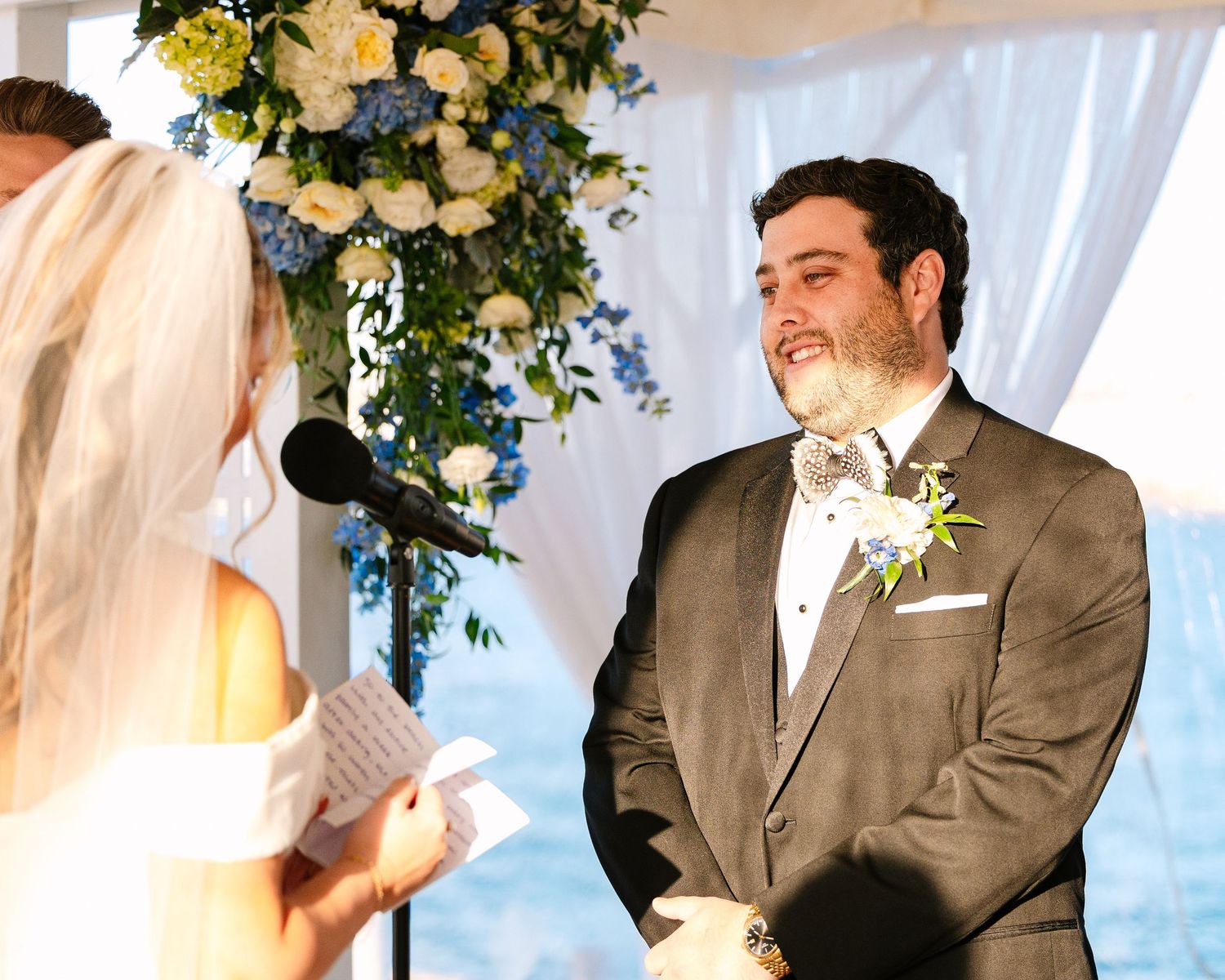 A couple exchanges vows during an indoor wedding ceremony under a floral arch with yellow and white flowers.