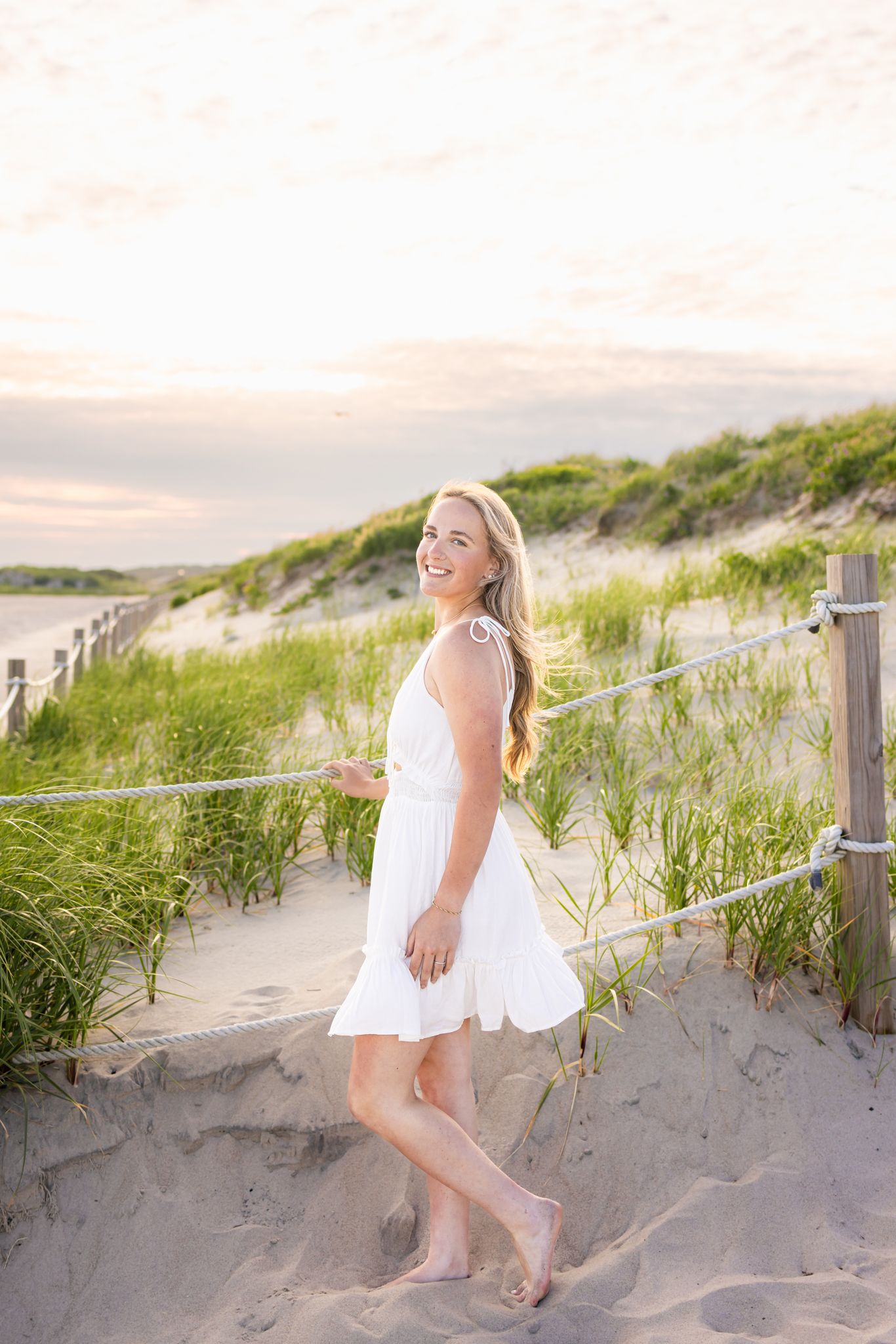 Cheerful blonde woman in white sundress posing on sandy beach path with dunes and grass.