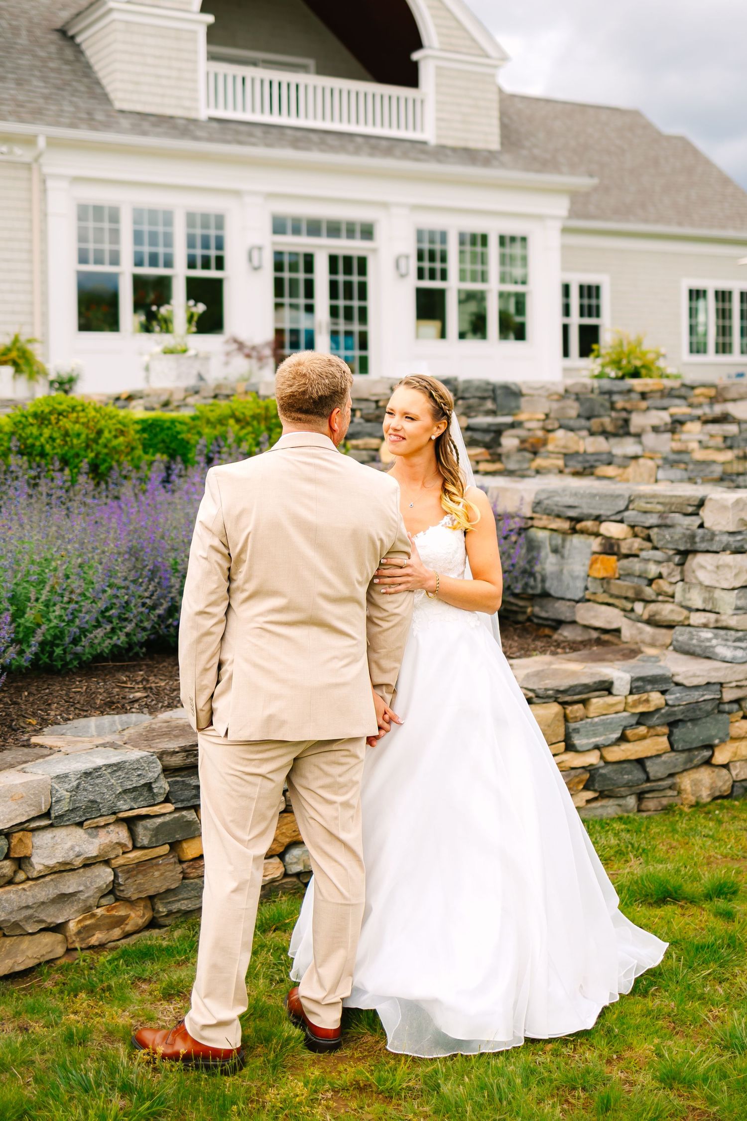 A couple in wedding attire stands on a lawn with stone wall and white house with balcony in the background.