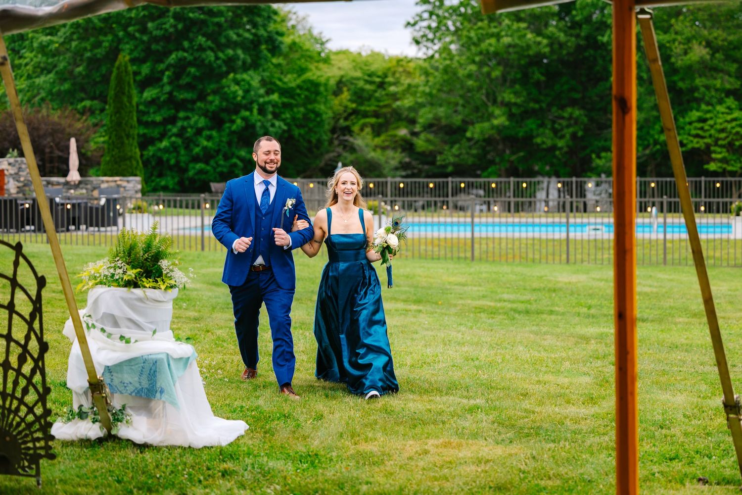 A couple stands together in a lush green garden setting with outdoor furniture and decor visible in the background.