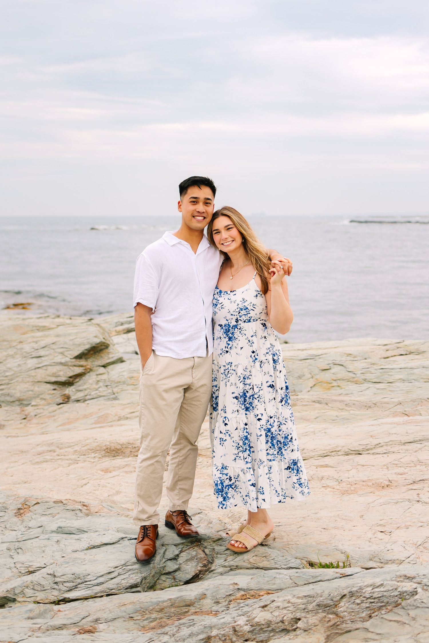 A couple poses together on rocky coastal terrain on a cloudy day after surprise proposal on Ocean Drive in Newport Rhode Island