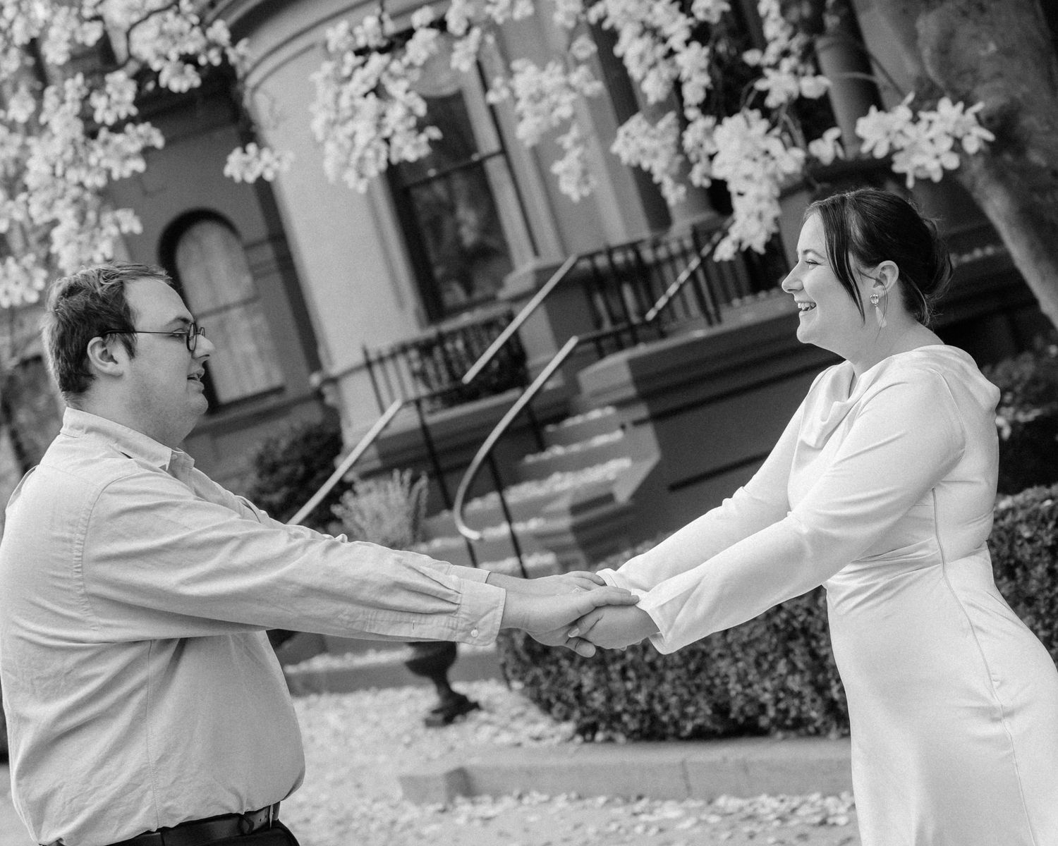 Black and white photo of a couple holding hands and smiling outdoors near a flowering tree.