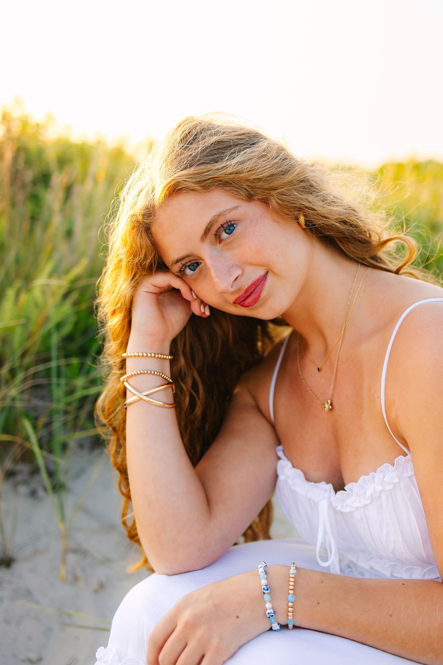 Close-up portrait of blue-eyed blonde woman in white dress with gold bracelets at sunset beach.