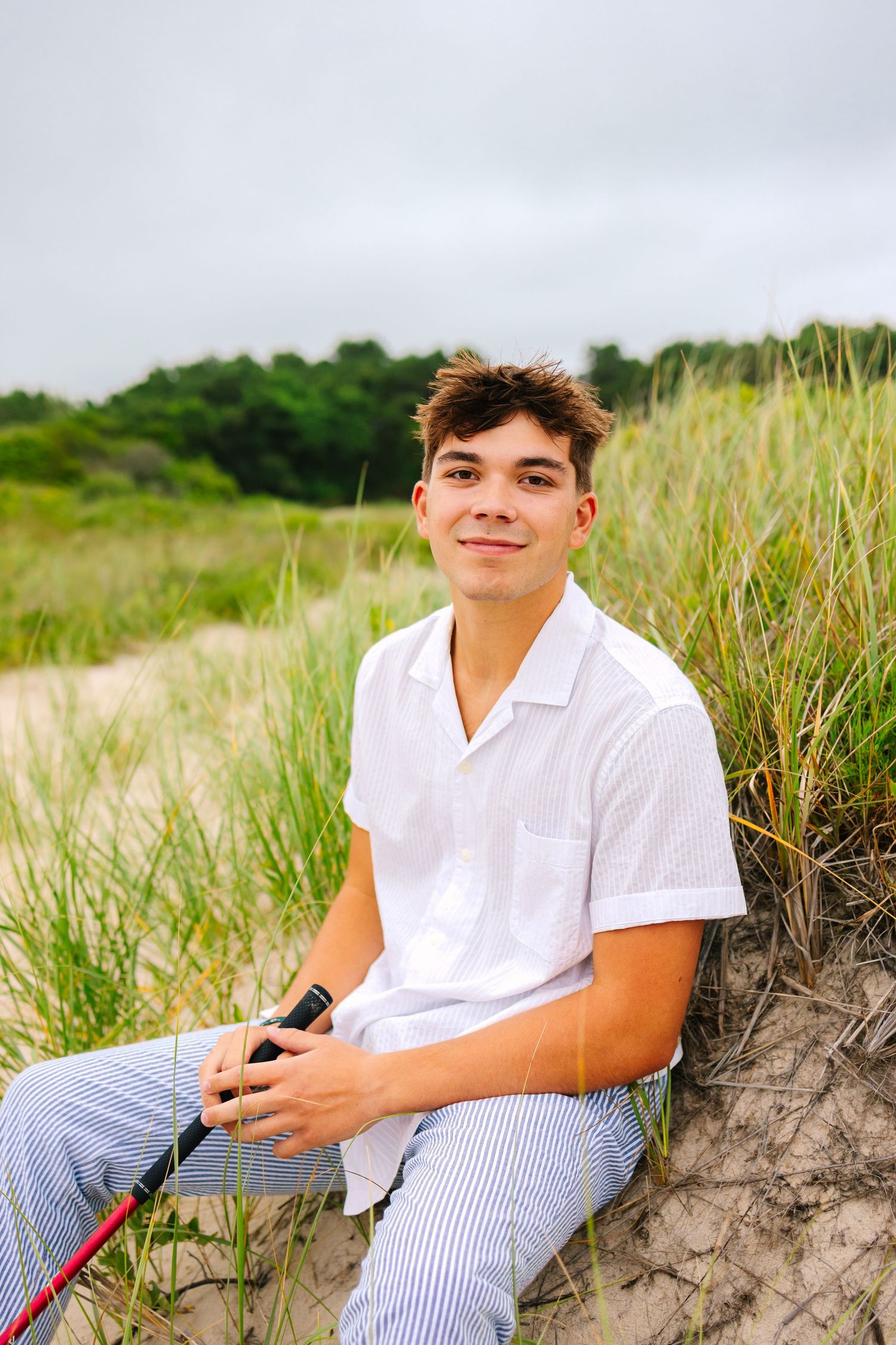 Smiling teenage boy in white shirt holding golf club on sandy beach dunes.