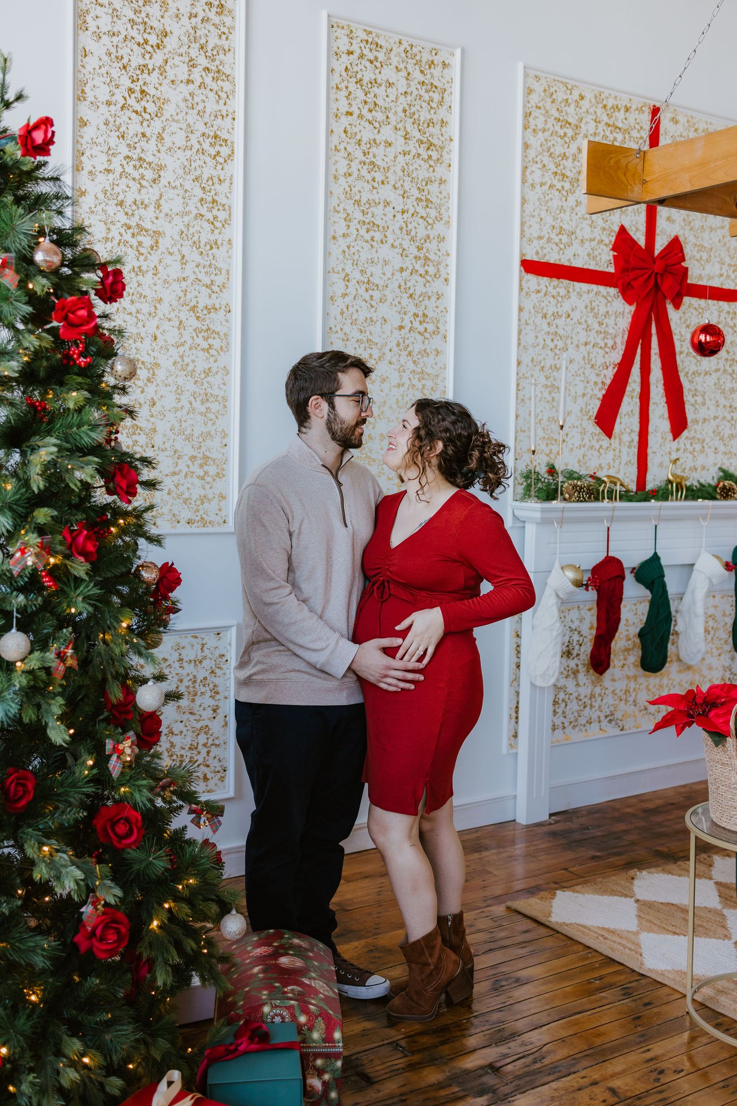 Couple embracing near a decorated Christmas tree, woman in red dress with visible baby bump.