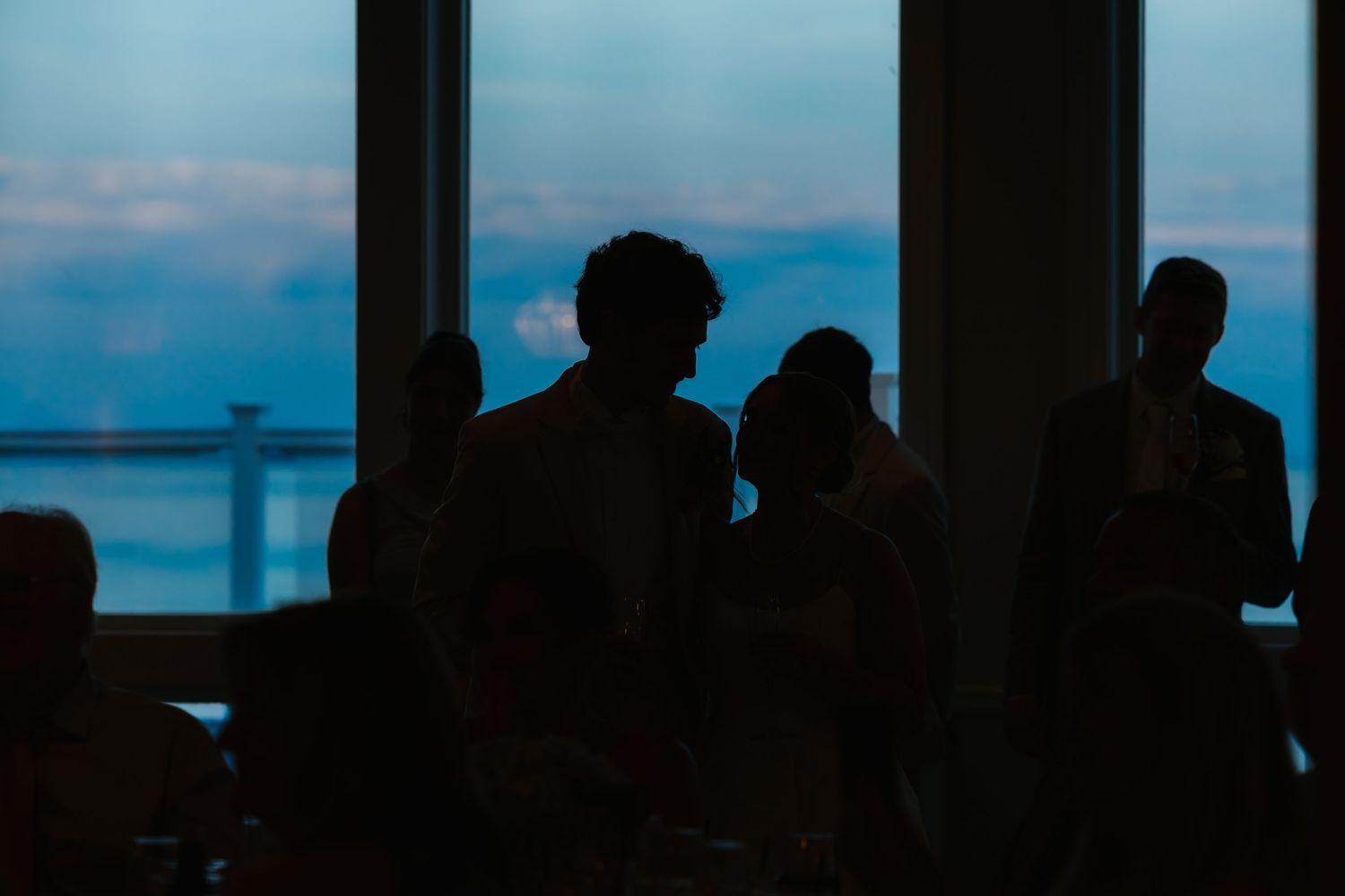 Silhouettes of wedding guests mingling near large windows with a stunning blue twilight sky backdrop.