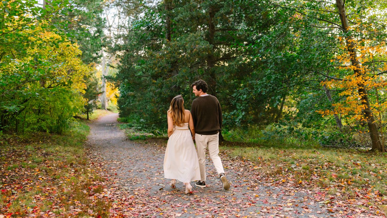 A romantic couple walks along a winding autumn path surrounded by lush green trees and golden foliage.
