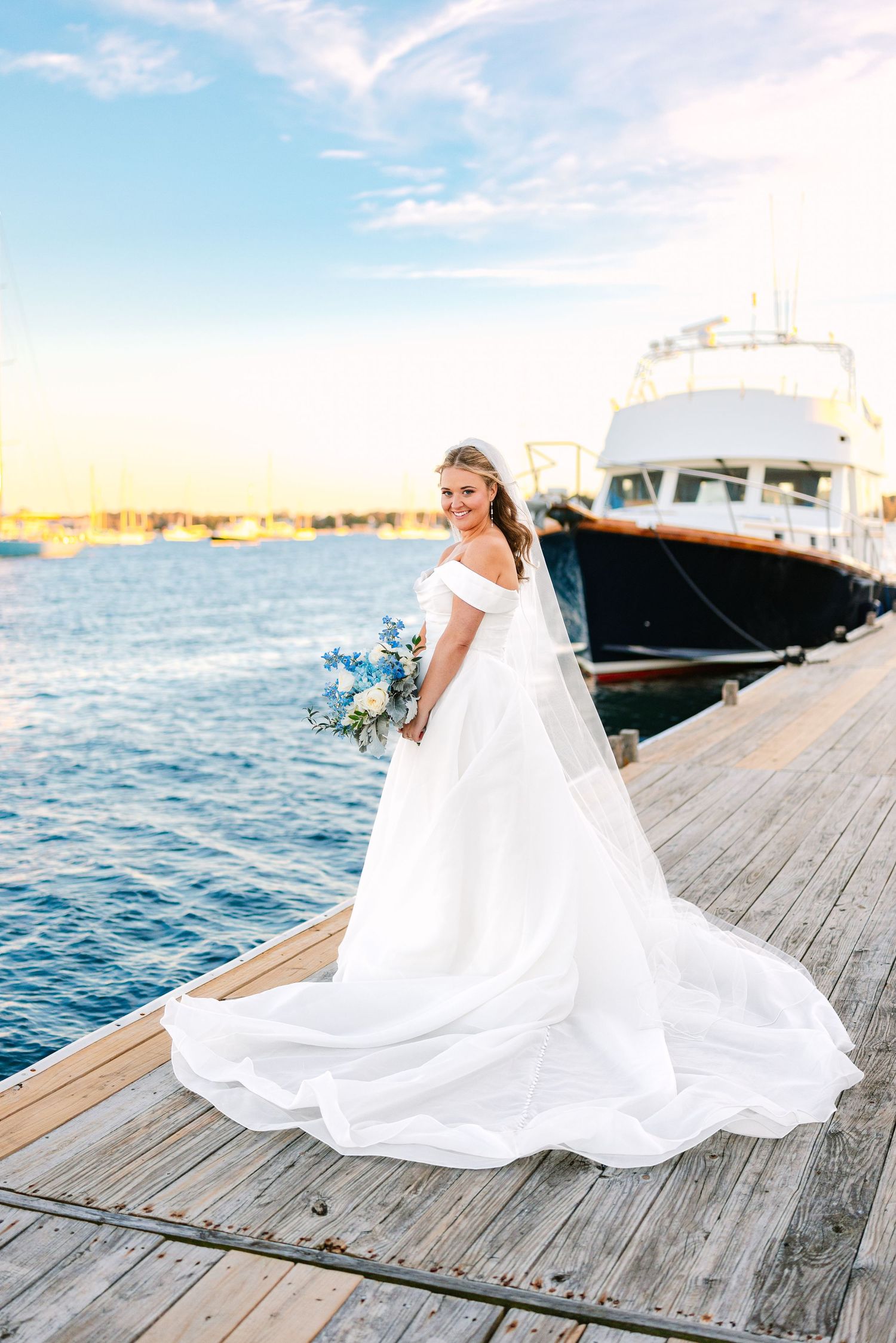 A bride in a flowing white gown poses gracefully on a wooden dock by the ocean at sunset at The Regatta Place in Newport Rhode Island