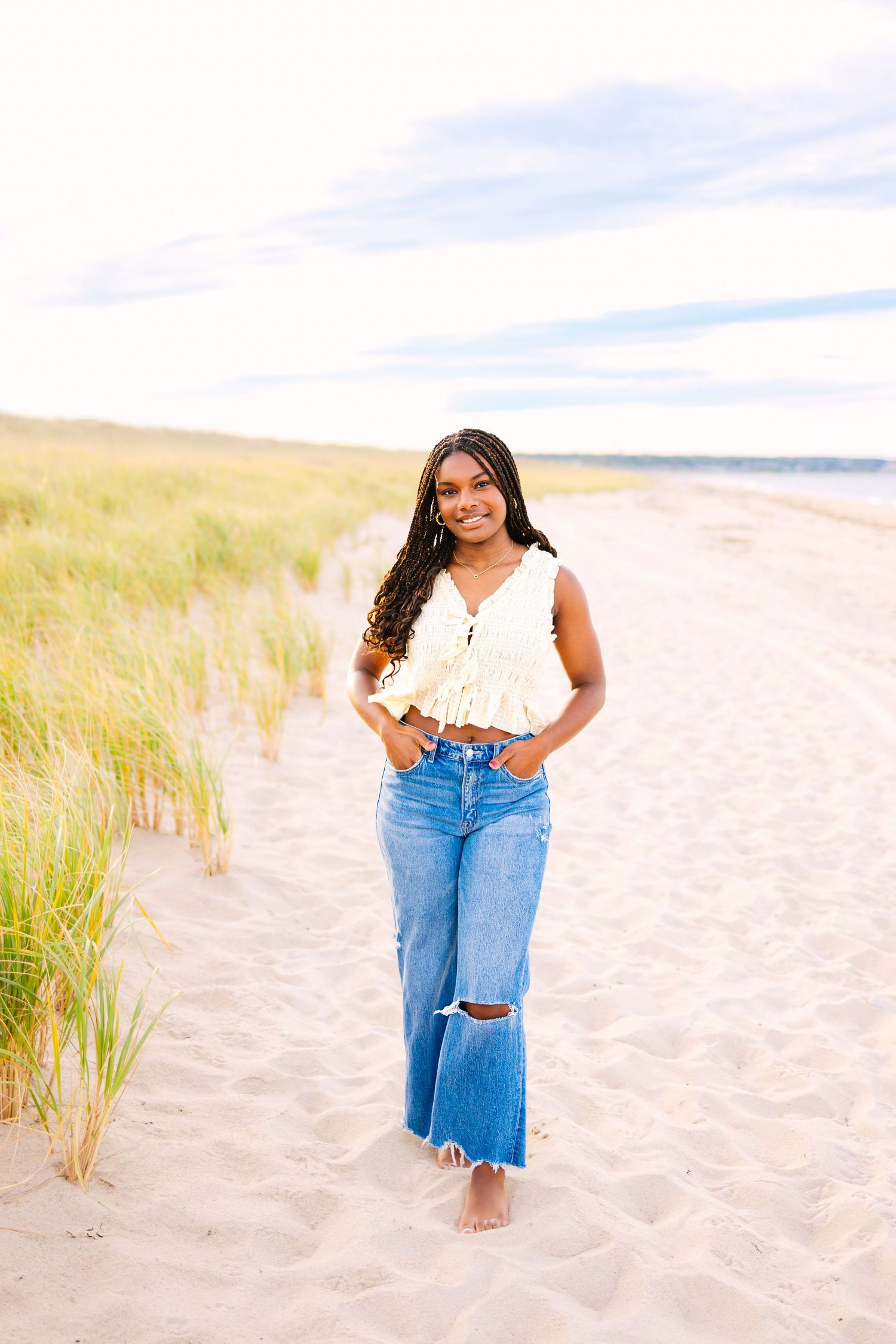 Young Black woman in white crop top and ripped jeans walking barefoot on sandy beach path with grass dunes.