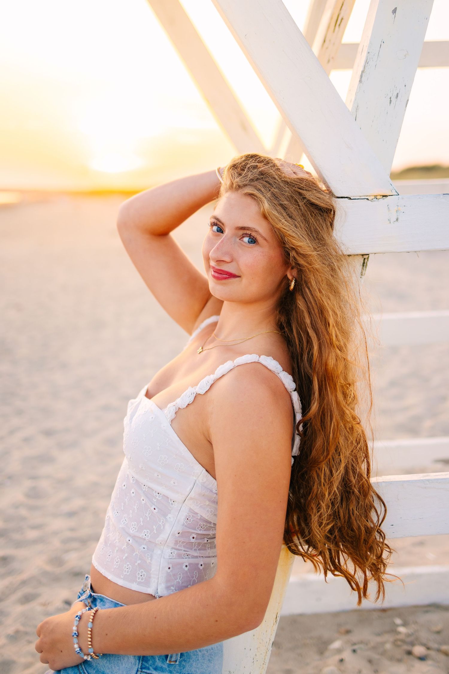 Young woman in white crop top and jeans leaning on lifeguard stand at sunset beach.