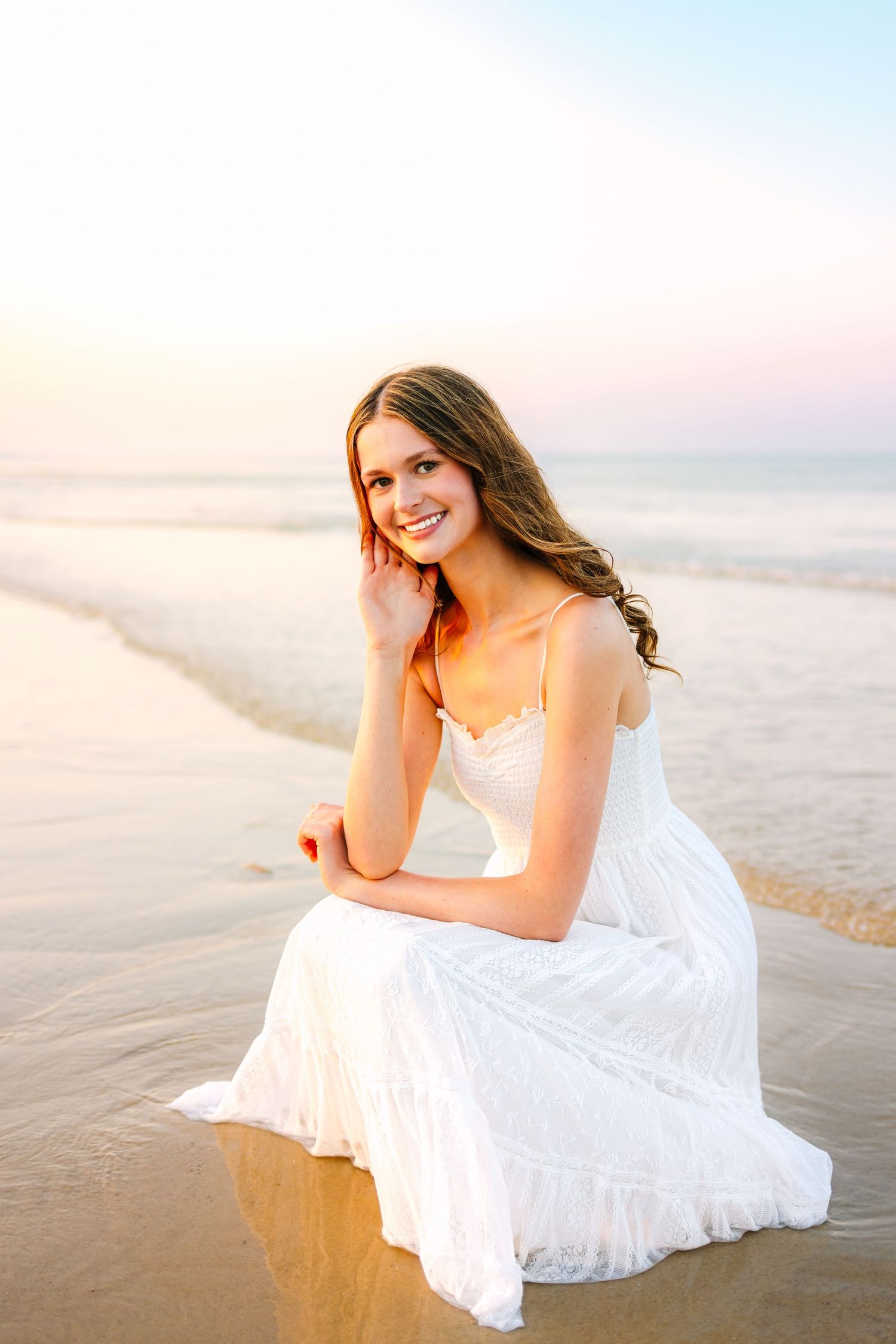 Young woman in white dress sitting on wet sand at beach, smiling warmly at golden sunset.