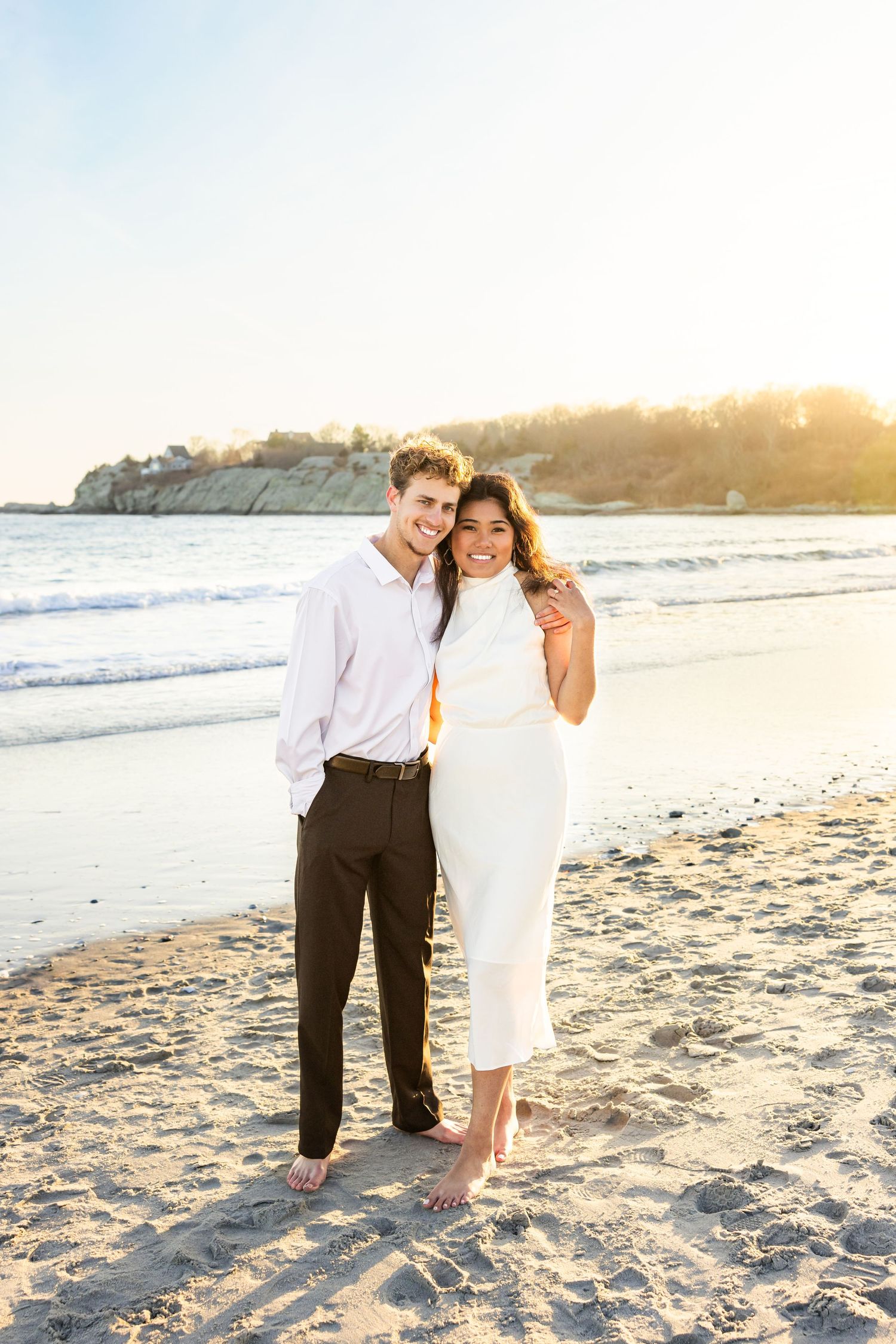 A couple stands together on a sandy beach at sunset with ocean waves and cliffs in the background.