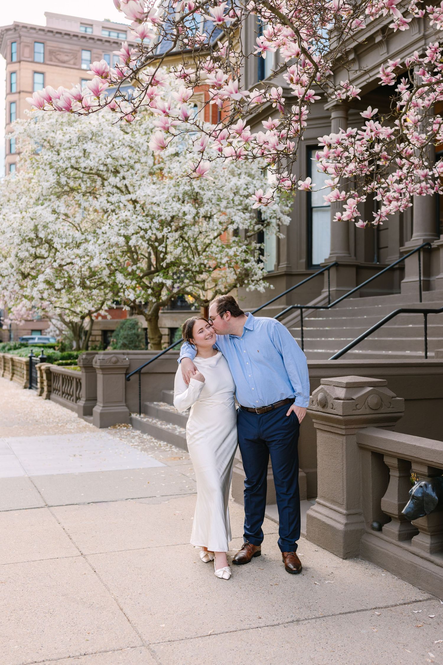 Couple sharing a tender kiss on a city sidewalk beneath blooming pink magnolia trees in spring.