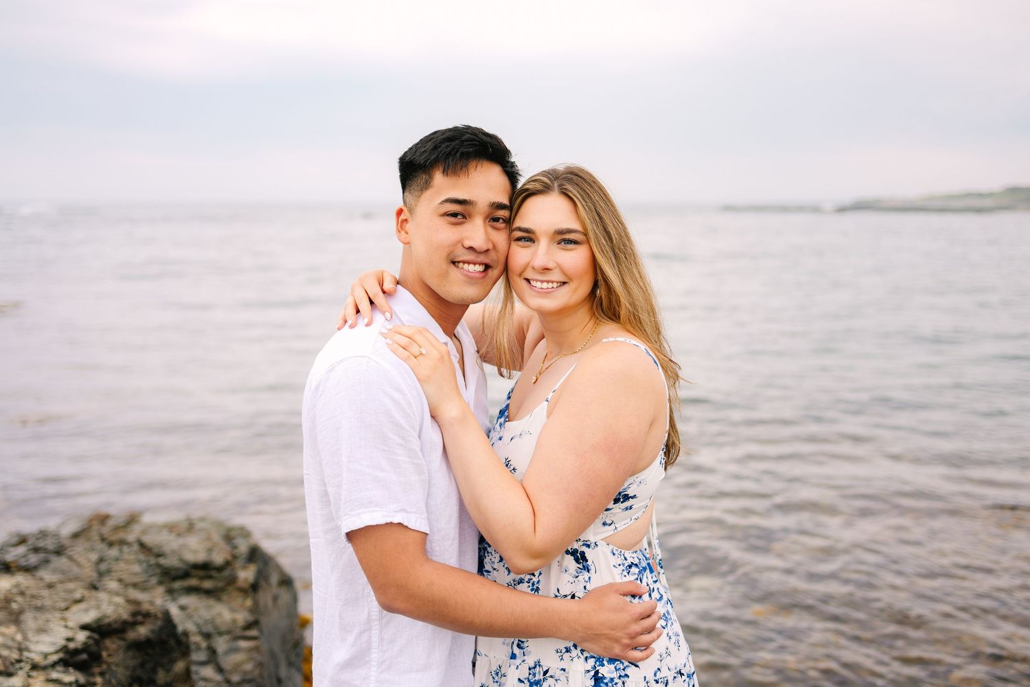 A newly engaged couple embraces and smiles at the camera on a rocky shoreline with ocean waves in the background in Newport Rhode Island.