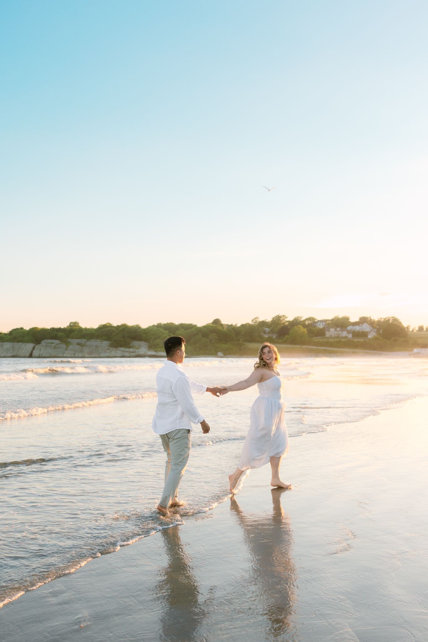 A couple in white outfits hold hands and walk along a sunlit beach at golden hour sunset.