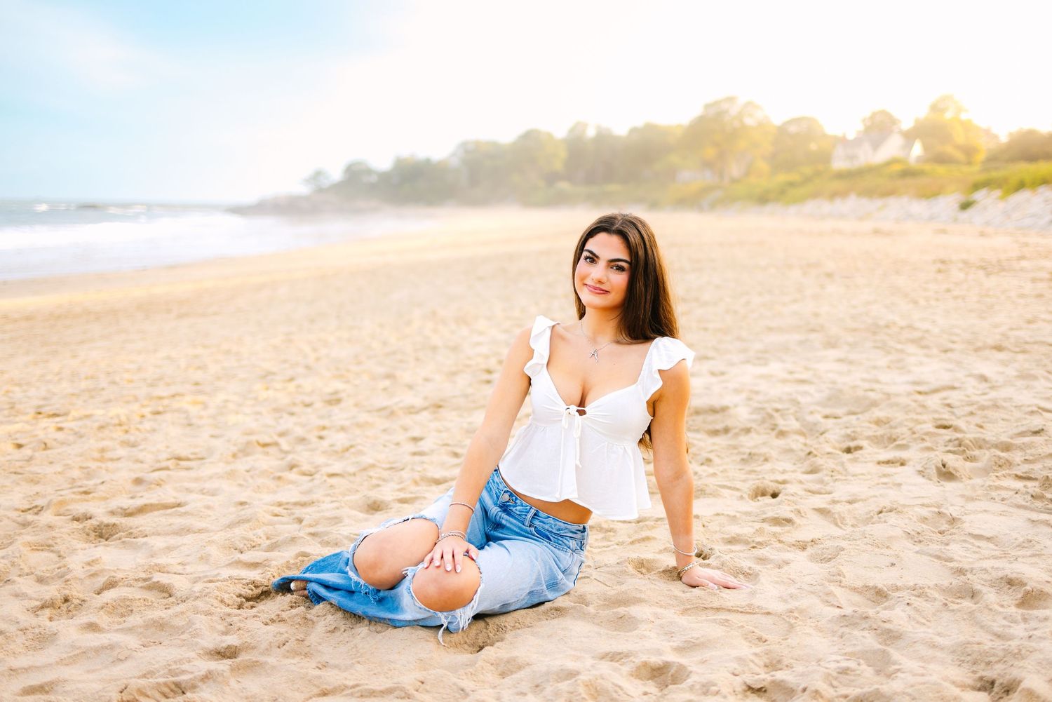 Young woman in white top and ripped jeans sitting relaxed on sandy beach in golden sunlight.