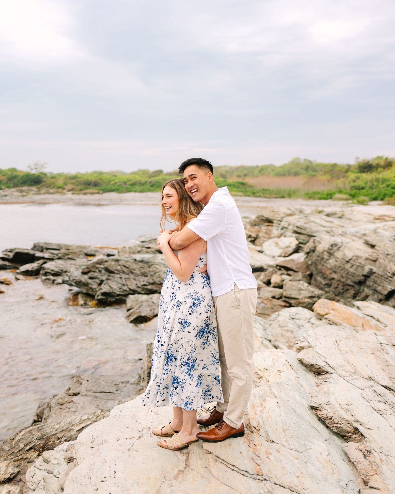 Couple embracing on rocky coastal shore for engagement photos, woman in floral dress, man in white shirt, smiling on Ocean Drive in Newport